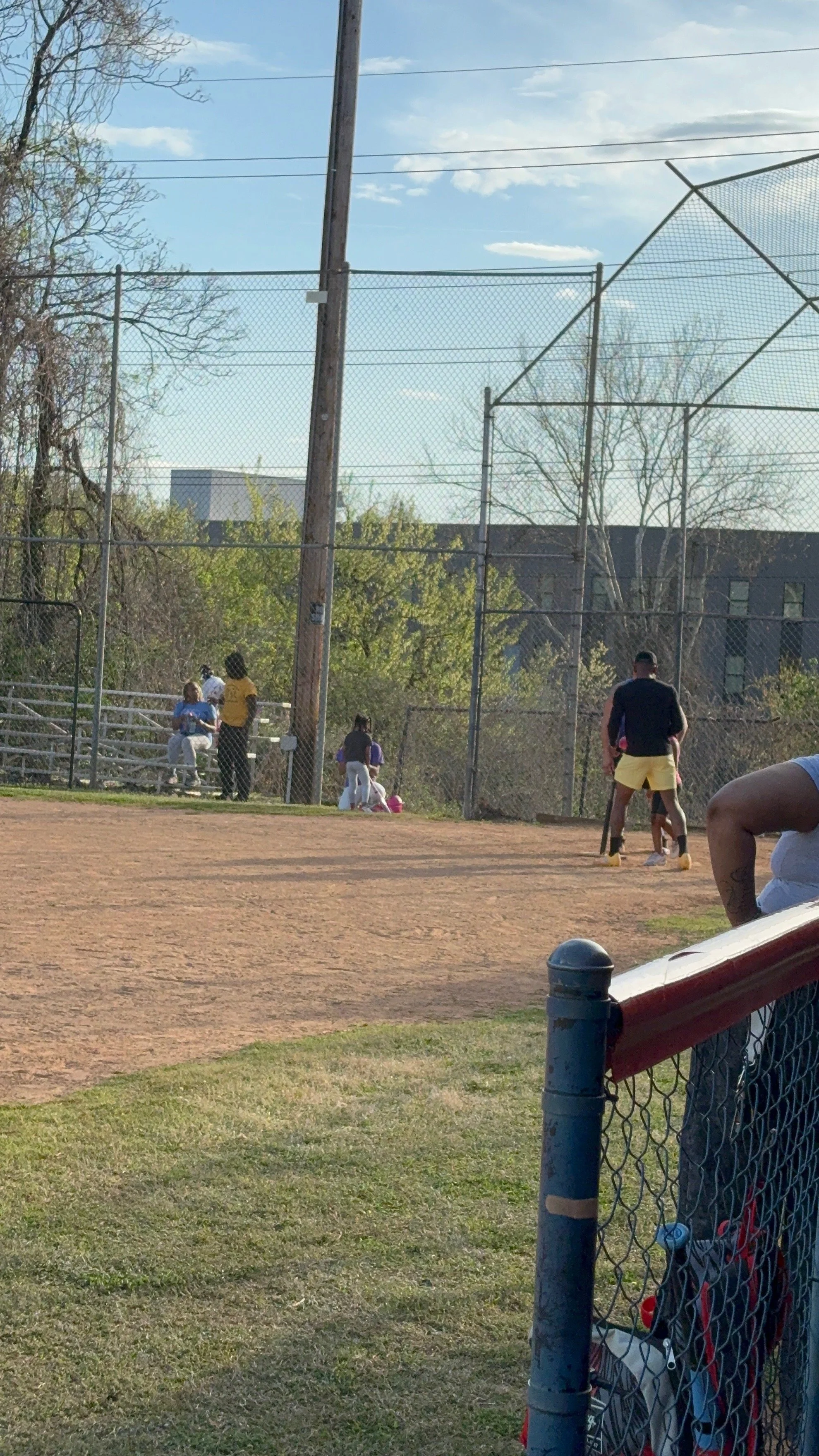 People at a baseball field, some sitting on bleachers and some standing near the fence, with a dirt field in the foreground and trees and buildings in the background.