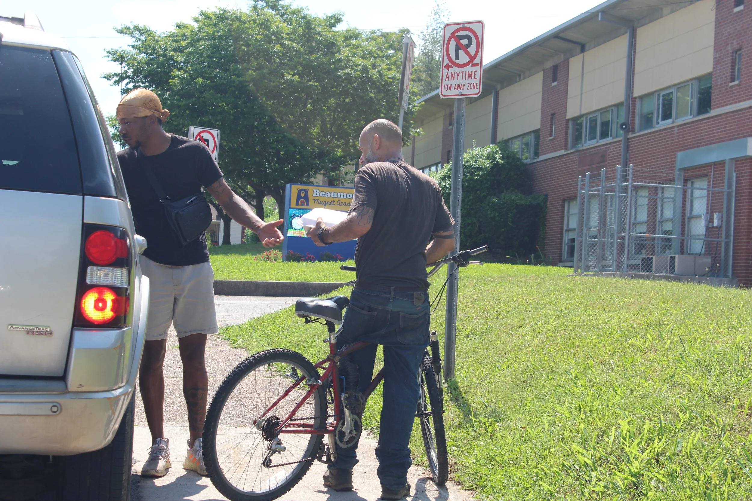 Two men stand near a car and bicycle on a sidewalk adjacent to a grassy area. One man is gesturing with his hand, and the other is holding papers, with a bicycle leaning against a pole nearby. Signage indicating no parking and tow-away zone is visibl