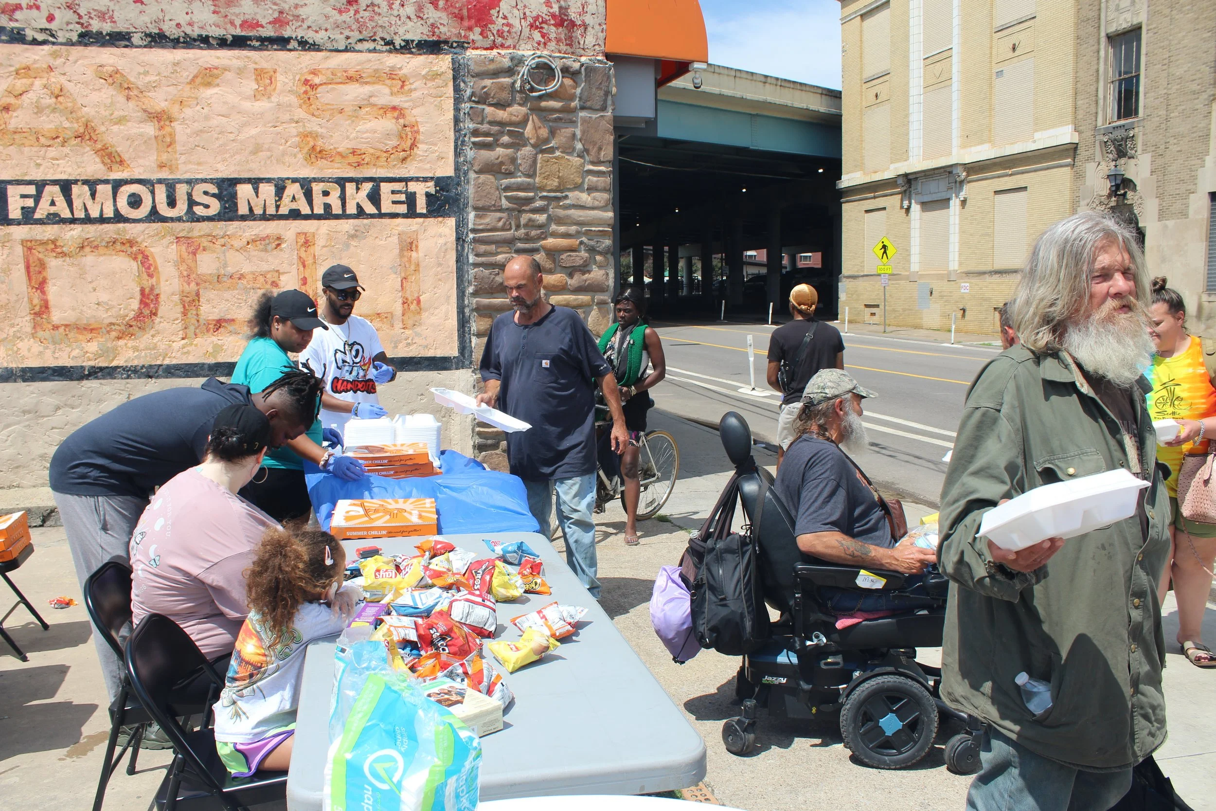People gather around a table with food and supplies outside a building near a street, possibly distributing aid or meals.