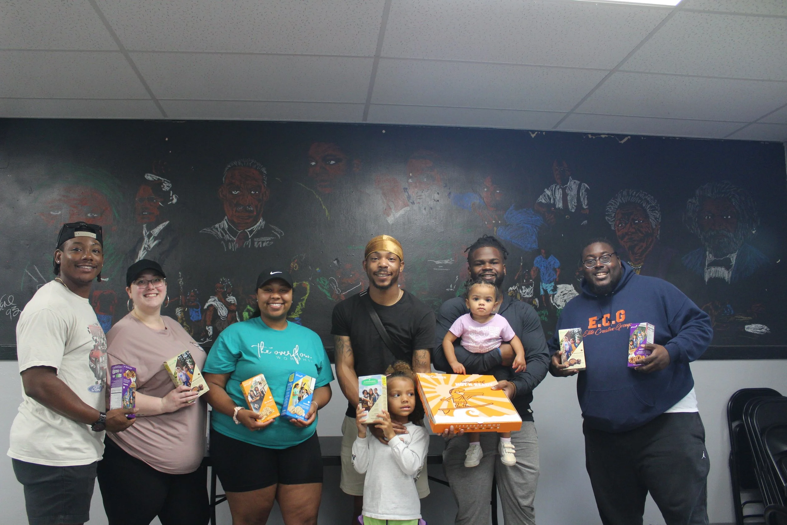 Group of seven people, including two children, holding food boxes and standing together in front of a black chalkboard wall with colorful chalk portraits, in an indoor setting.
