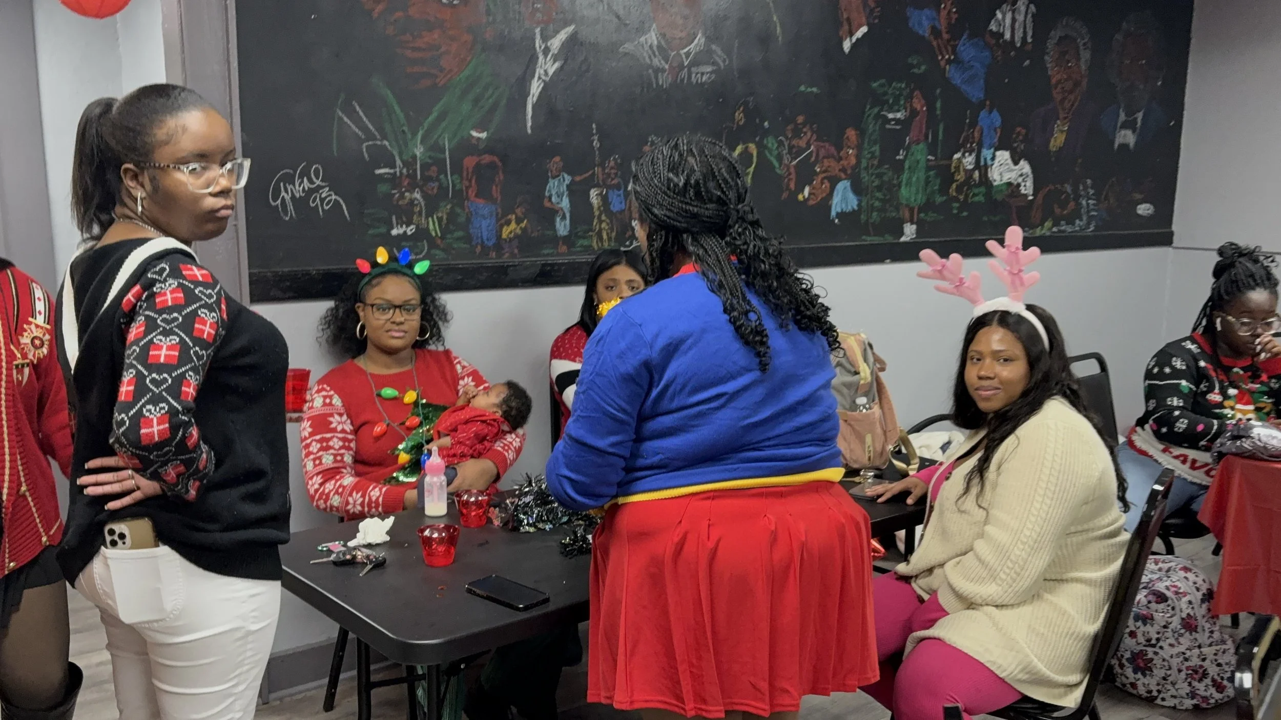Group of women celebrating Christmas indoors. Some women are wearing festive accessories like reindeer antlers and a Christmas light headband. One woman is holding a baby, and others are sitting or standing around a table with holiday decorations and