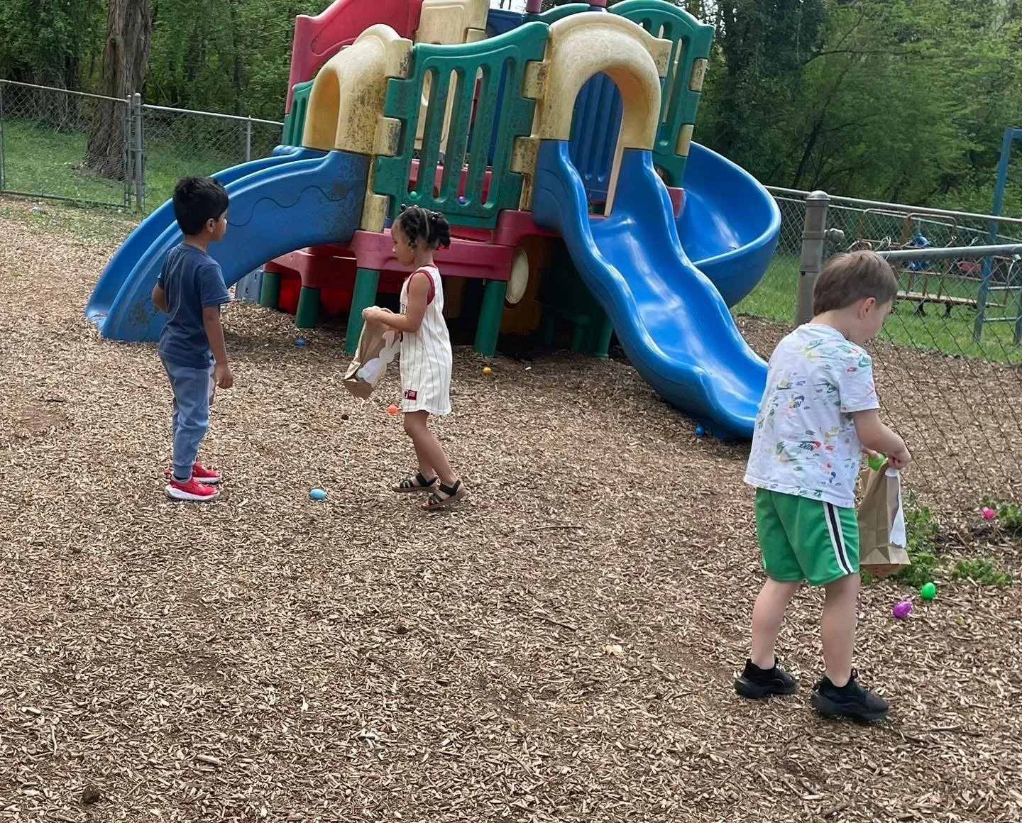 Three children at a playground near a blue slide, holding small bags and colorful plastic Easter eggs on the ground, with a forest background.