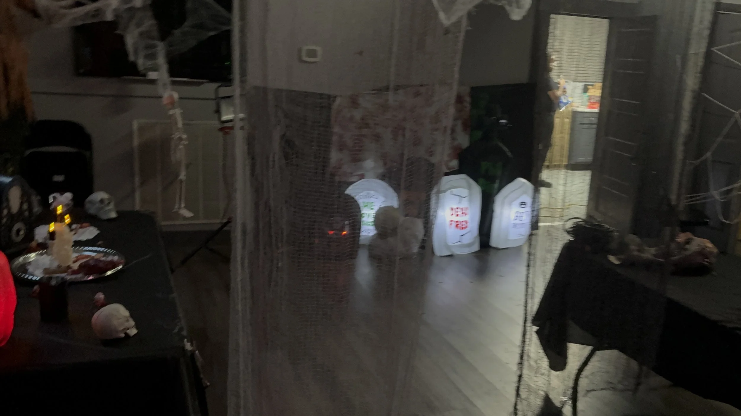 A dark room decorated for Halloween with a spooky table setting featuring skulls and candles, pumpkins, and a Halloween net draped over the scene. There are also some bags and boxes on the floor, and a person visible in the background near a kitchen 