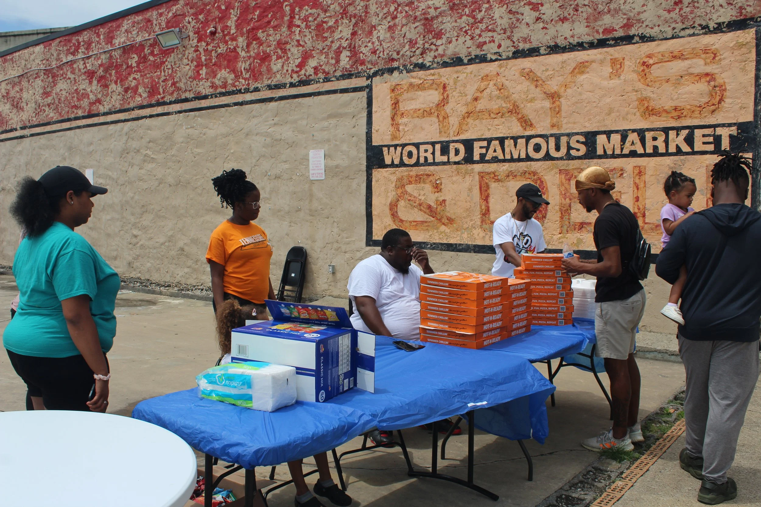 People gathered around tables selling pizza in front of a wall with a painted sign that reads 'Ray's World Famous Market & Store'.