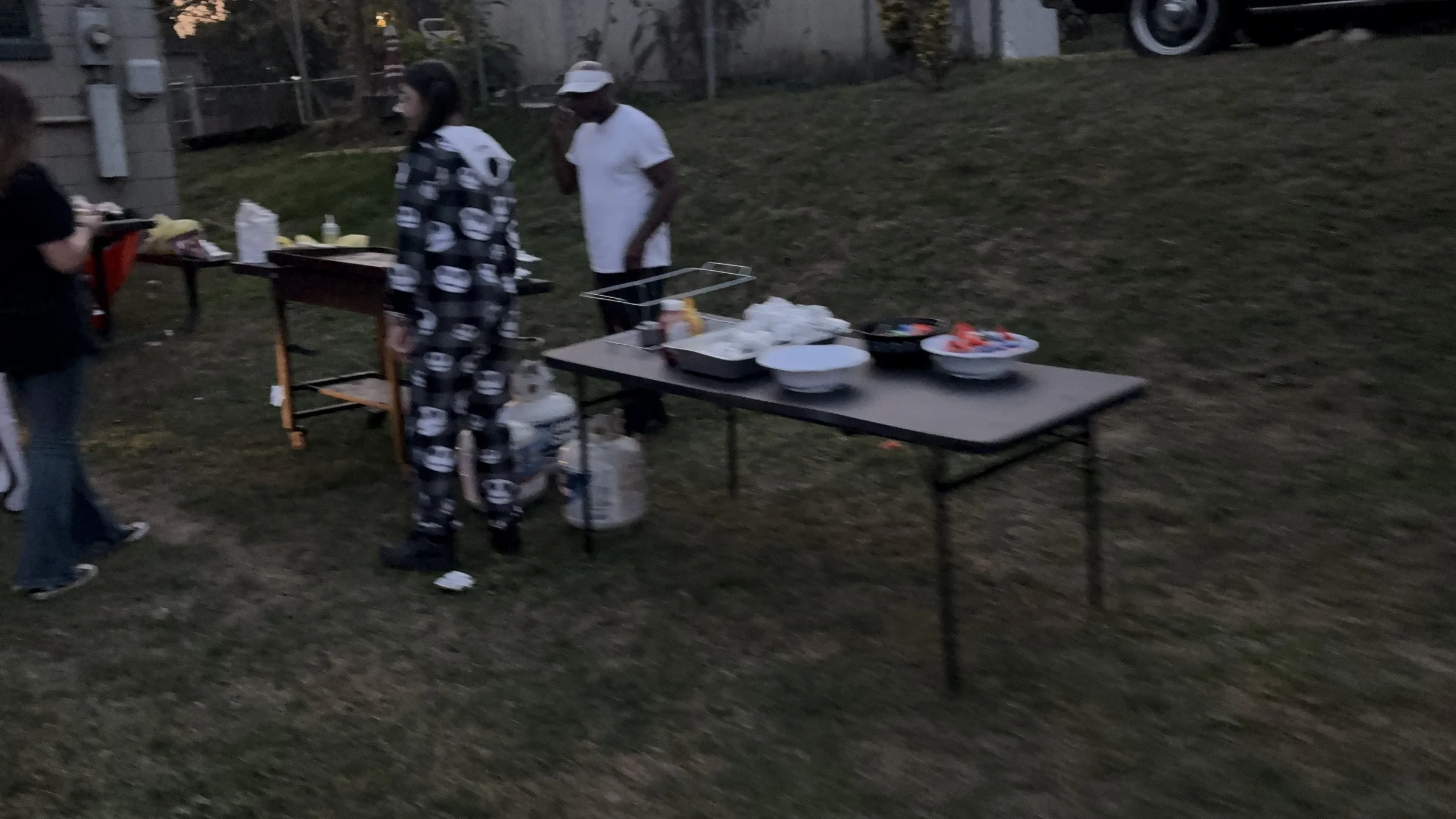 People at an outdoor gathering with a table of food, in a grassy area near a building, with four individuals visible, one in pajamas, another wearing a white hat and white shirt.