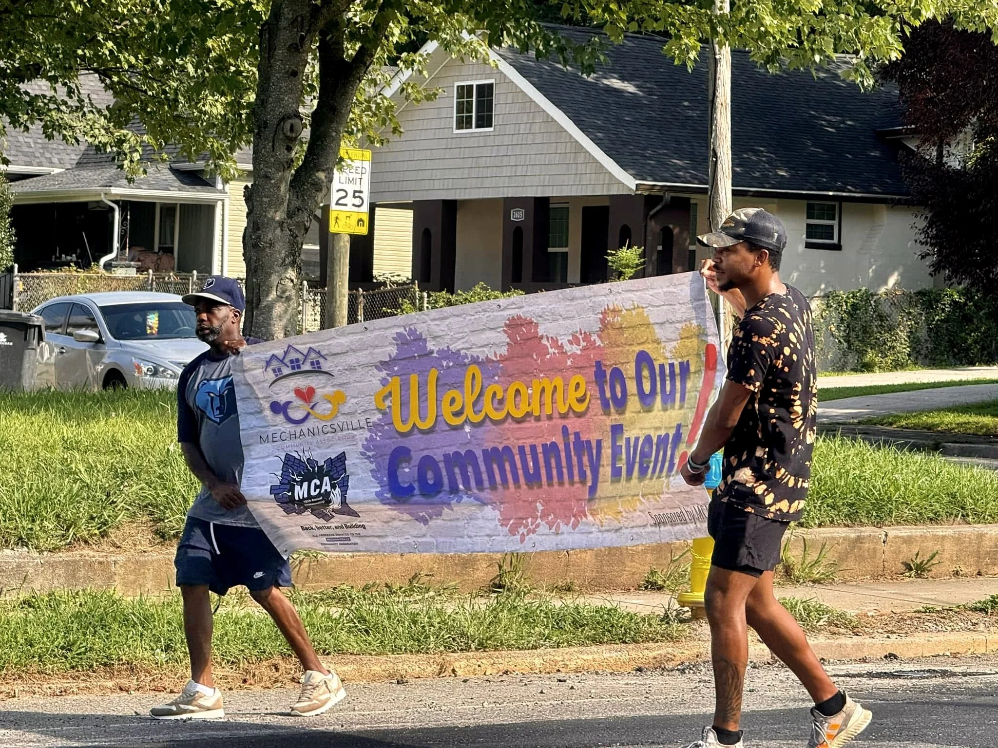 Two men are walking on a street holding a colorful banner that reads, 'Welcome to Our Community Event!' The background features a residential neighborhood with homes, trees, and parked cars.
