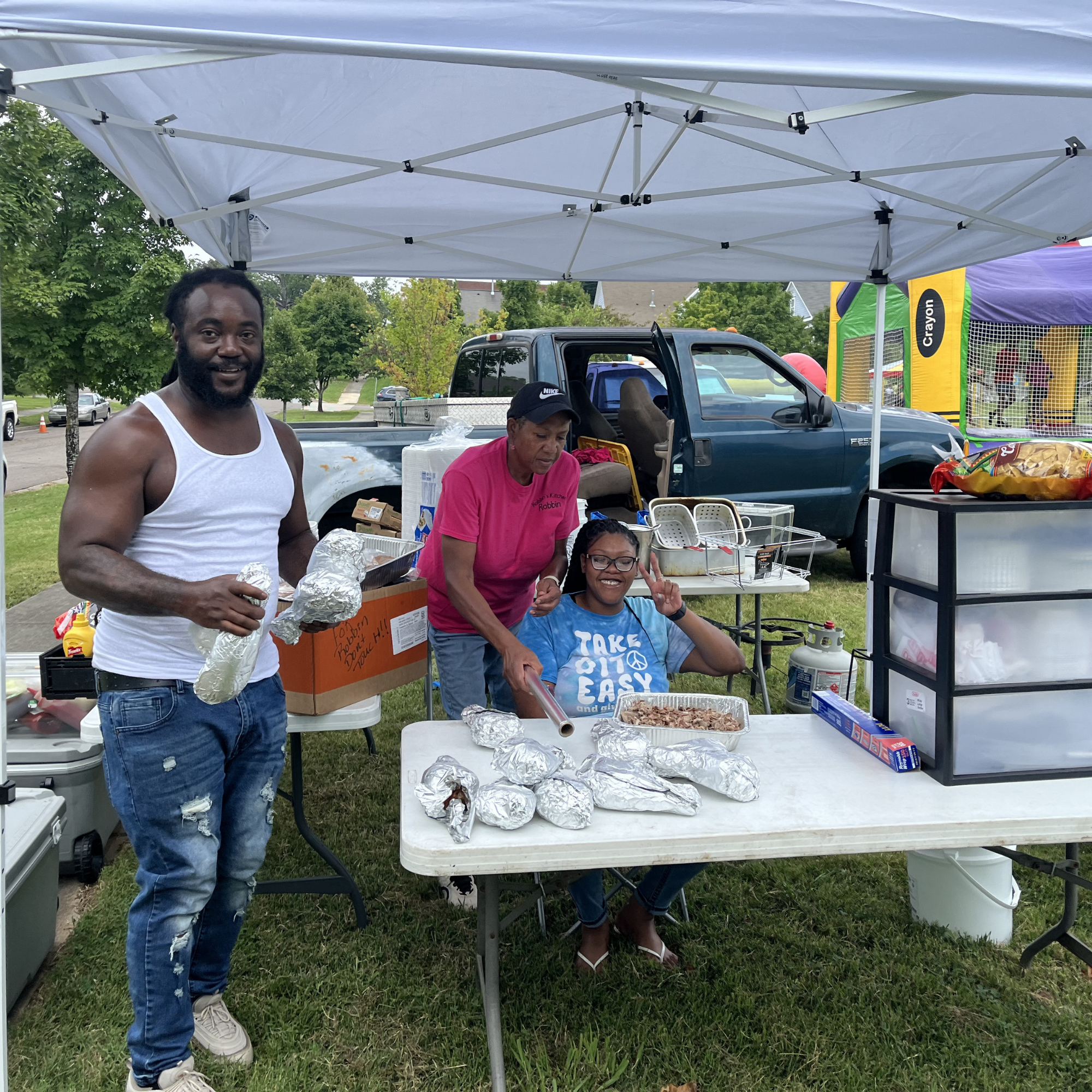 Three people standing and sitting at a cookout table with foil-wrapped food, with a pickup truck behind them and a bounce house in the background.