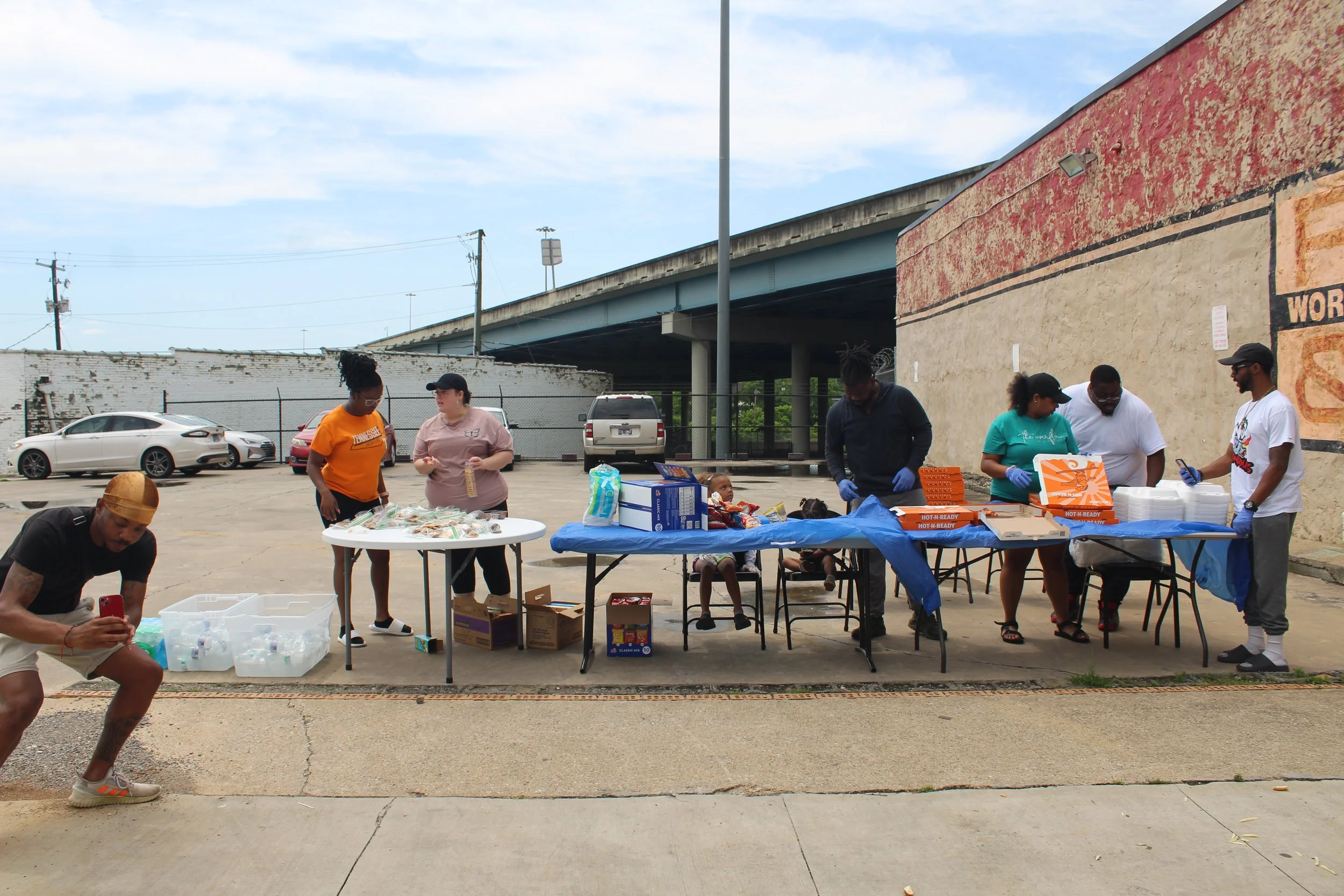 People organizing food and supplies at an outdoor setup, with tables filled with boxes and packages, under a highway overpass, in a parking lot.