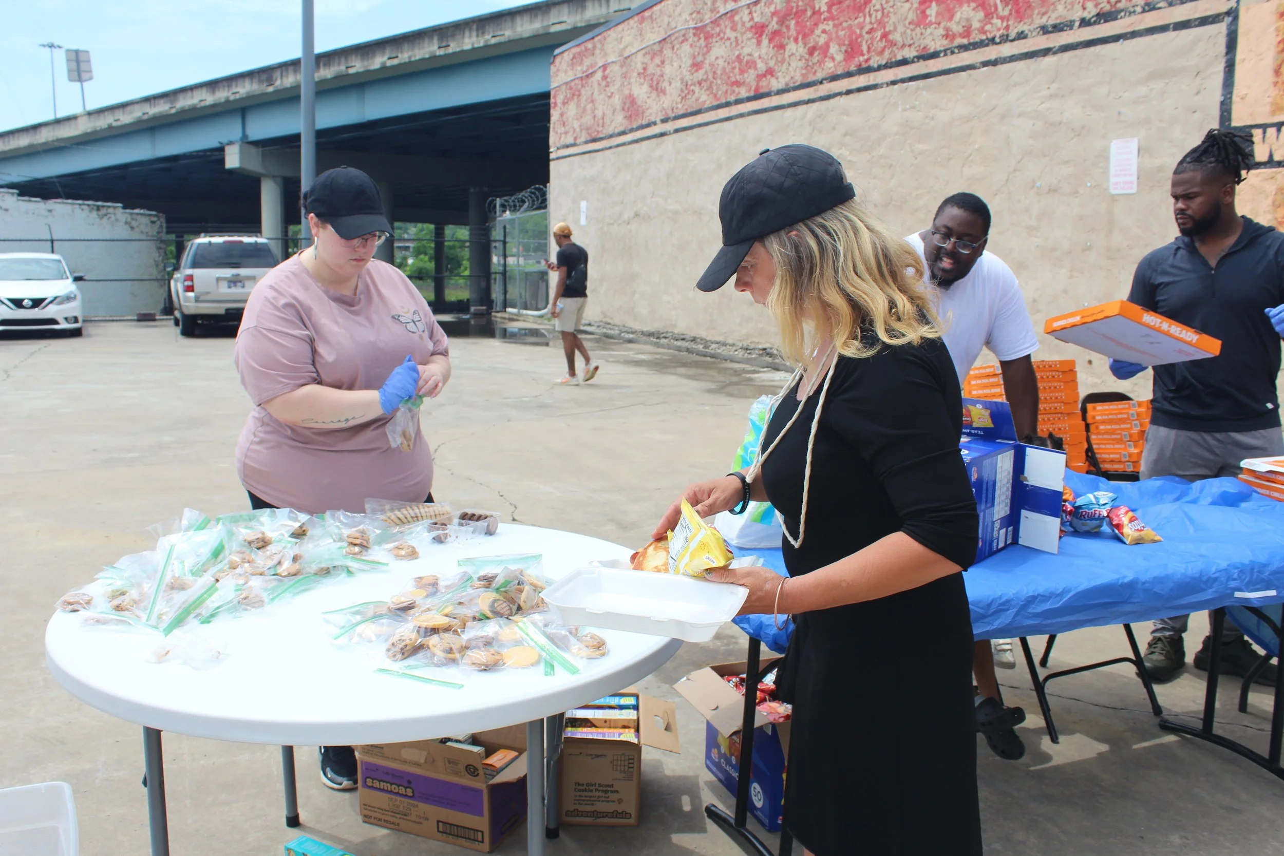 People distributing and receiving food and snacks at an outdoor event, with a table of individually bagged snacks and pizza boxes nearby.
