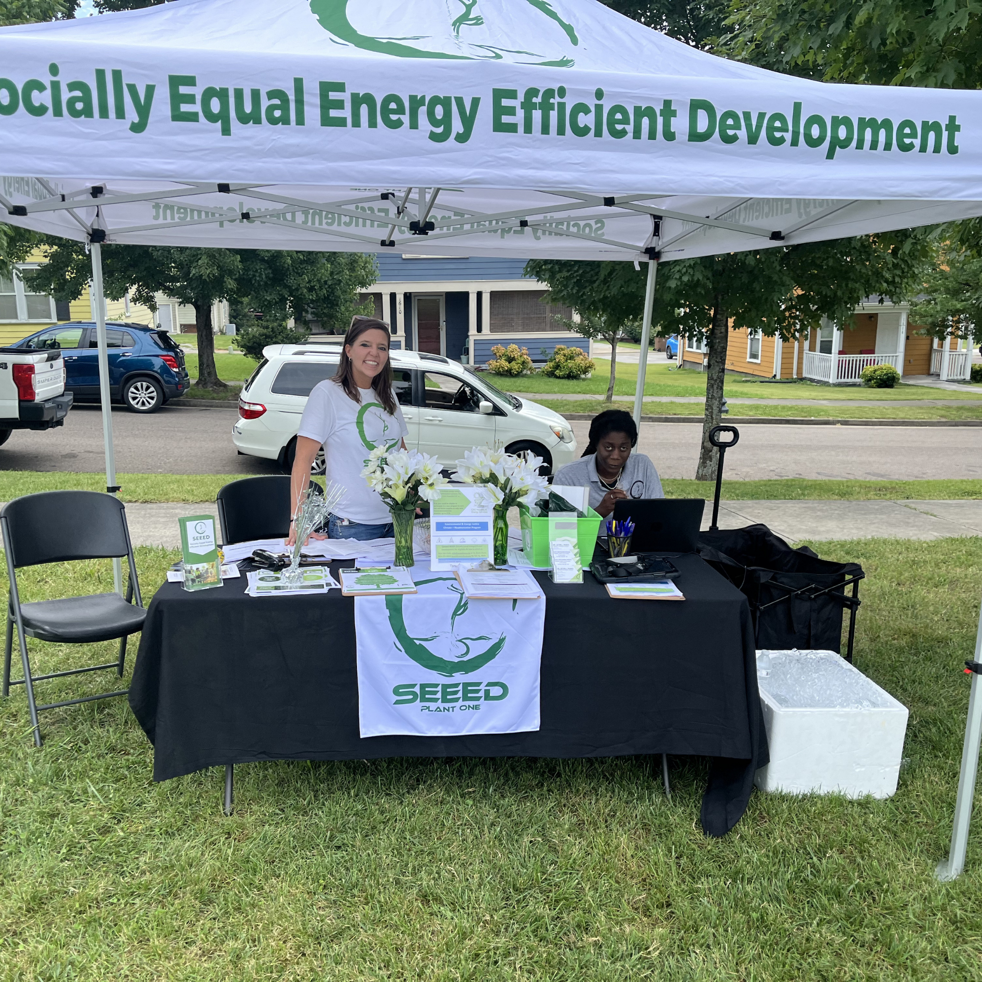 A booth set up outdoors under a white canopy tent with the text 'Socially Equal Energy Efficient Development' printed on it. The booth has a table covered with a black tablecloth displaying brochures, flowers, and informational materials. Two women a