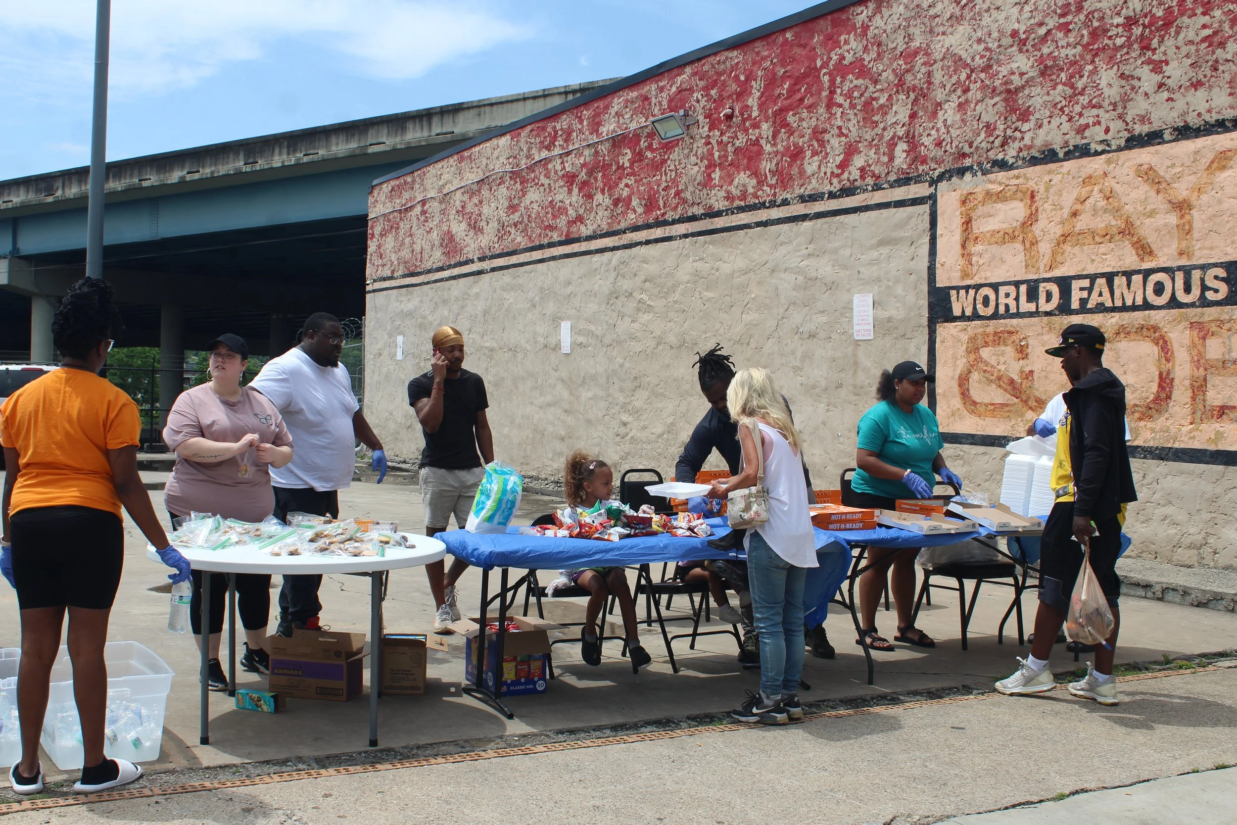 People gathered around tables outdoors, serving and receiving food and drinks, with a mural on the wall behind them.