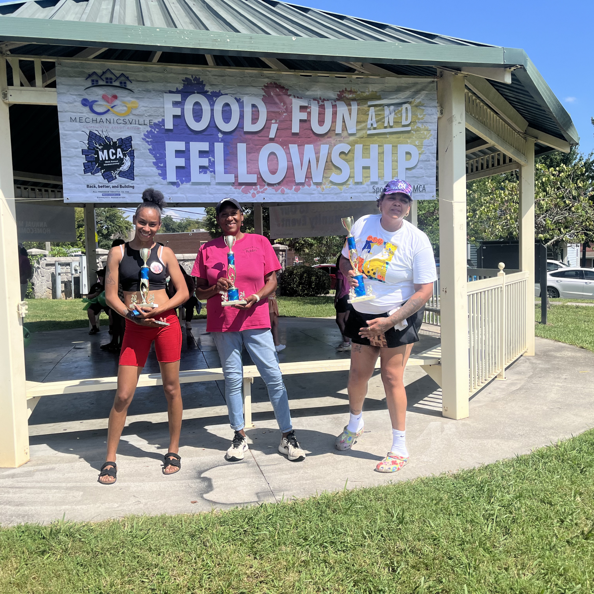 Three women standing on a stage under a banner that reads "Food, Fun, and Fellowship." Two women are holding trophies, and the woman on the right is holding a trophy and wearing a colorful cap. All are smiling with a sunny outdoor setting in the back