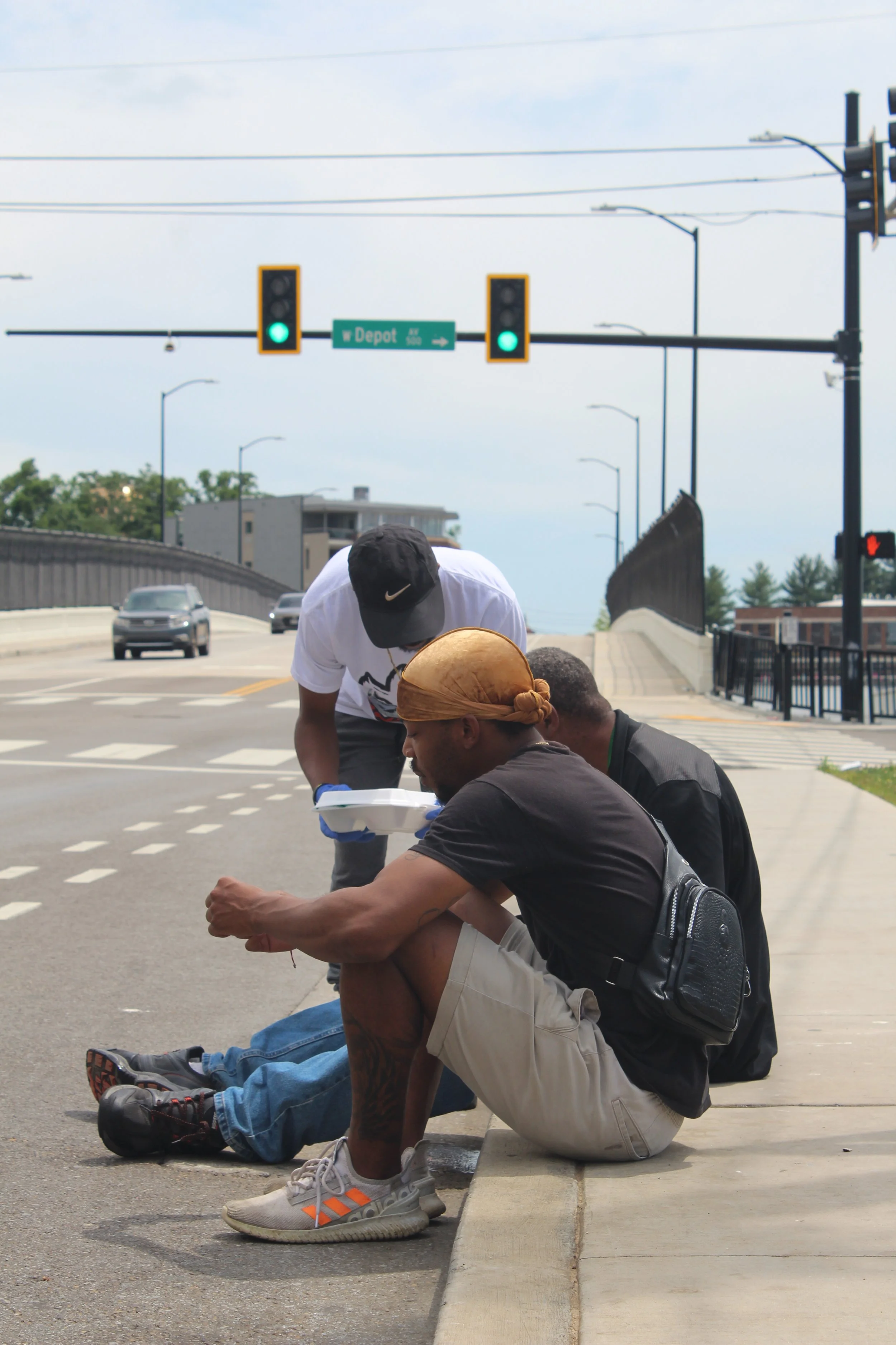 Three men sitting and standing on a sidewalk near a street with green traffic lights, holding food containers.