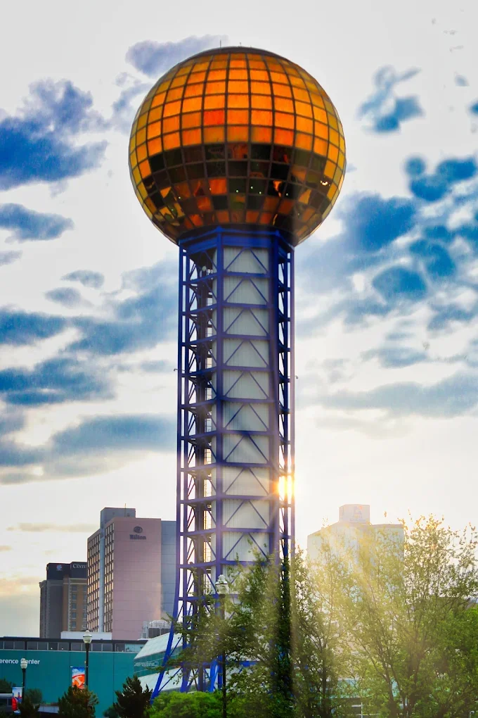 A tall tower with a large, spherical, gold-colored top, with a lattice-like blue and white support structure, set against a partly cloudy sky at sunset.