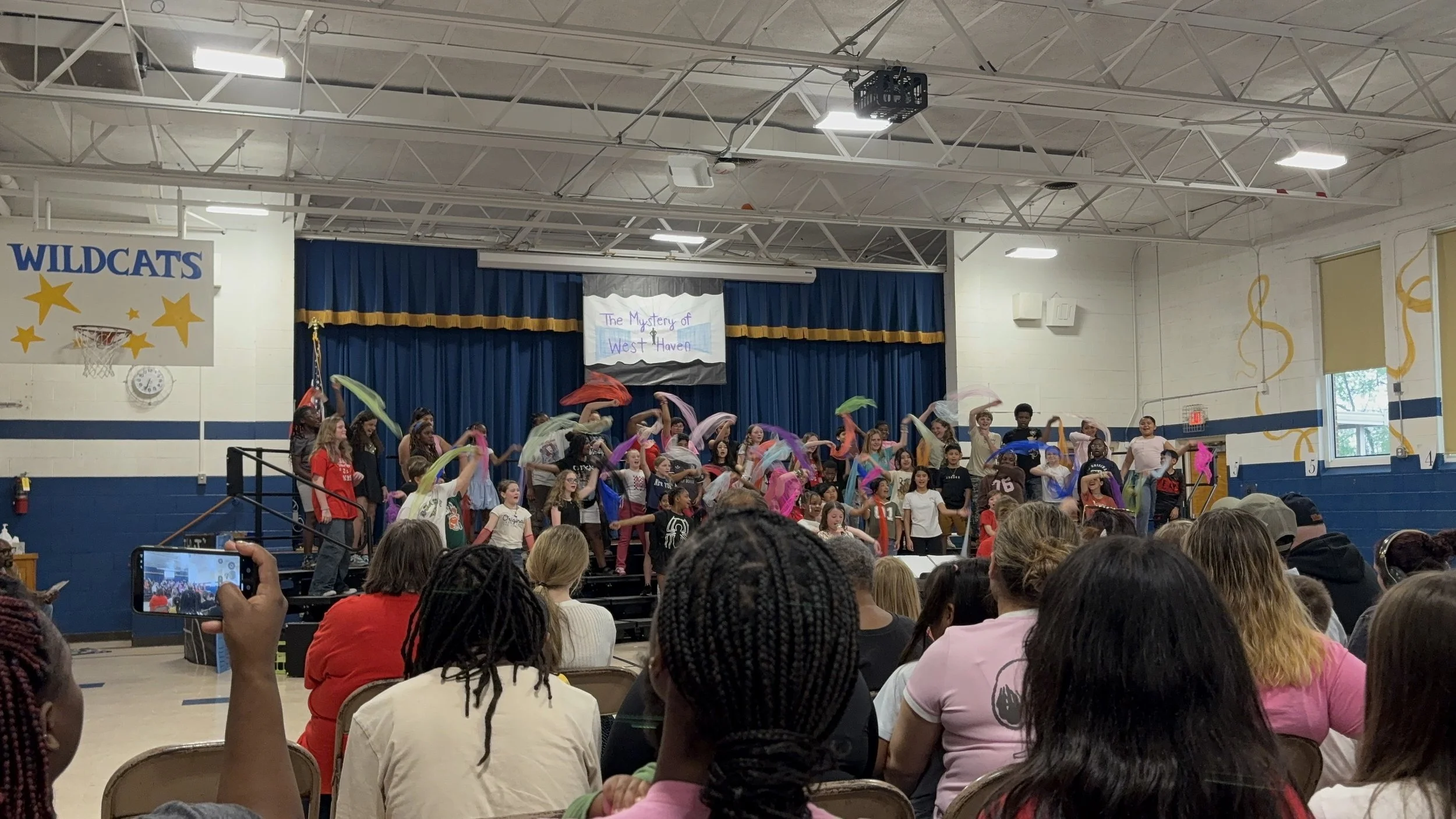 Children performing a dance on stage during a school event titled "The Mystery of West Haven" with an audience watching, some recording.