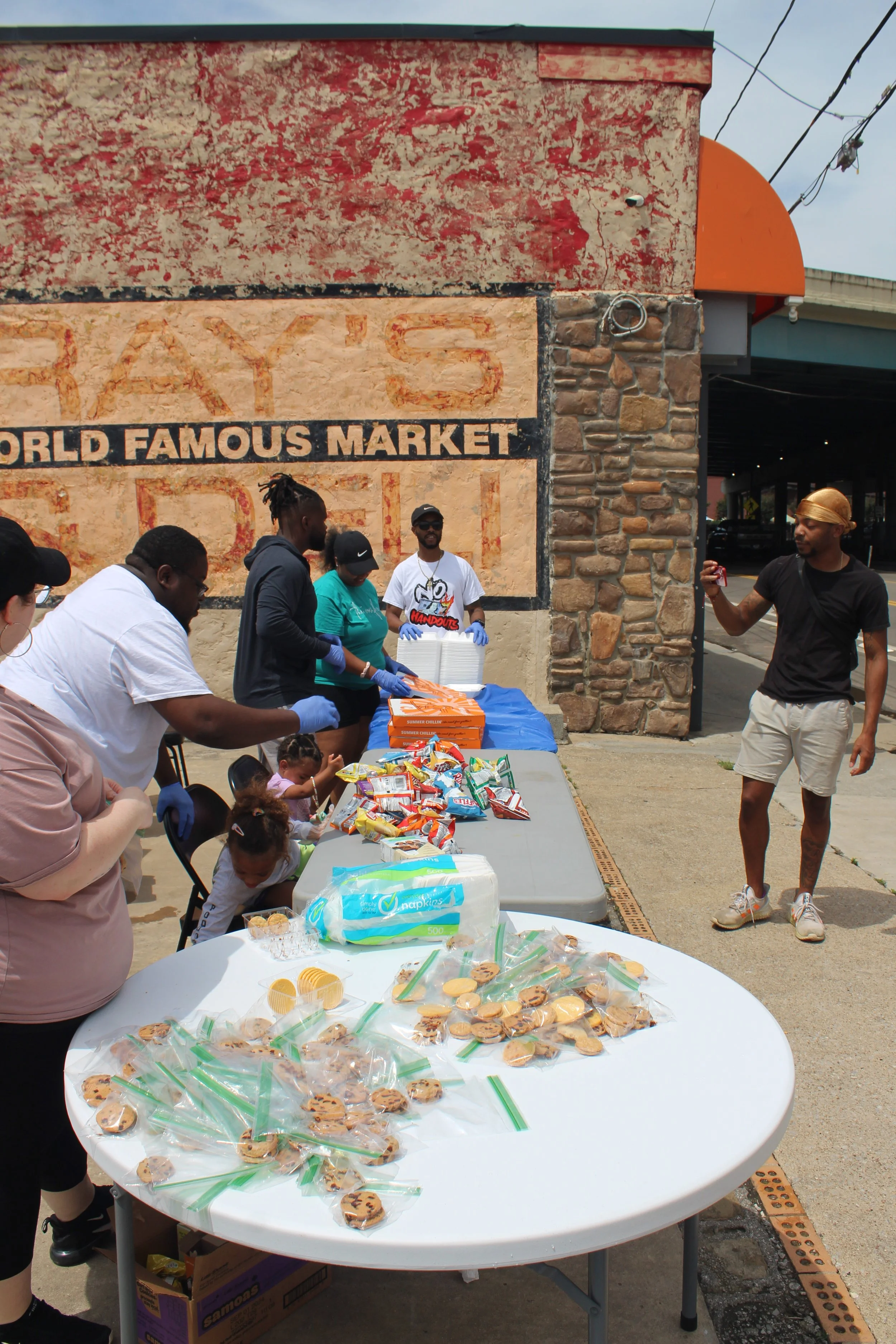 People serving and receiving snacks outside a market, with a wall that says 'FAMOUS MARKET' in the background.