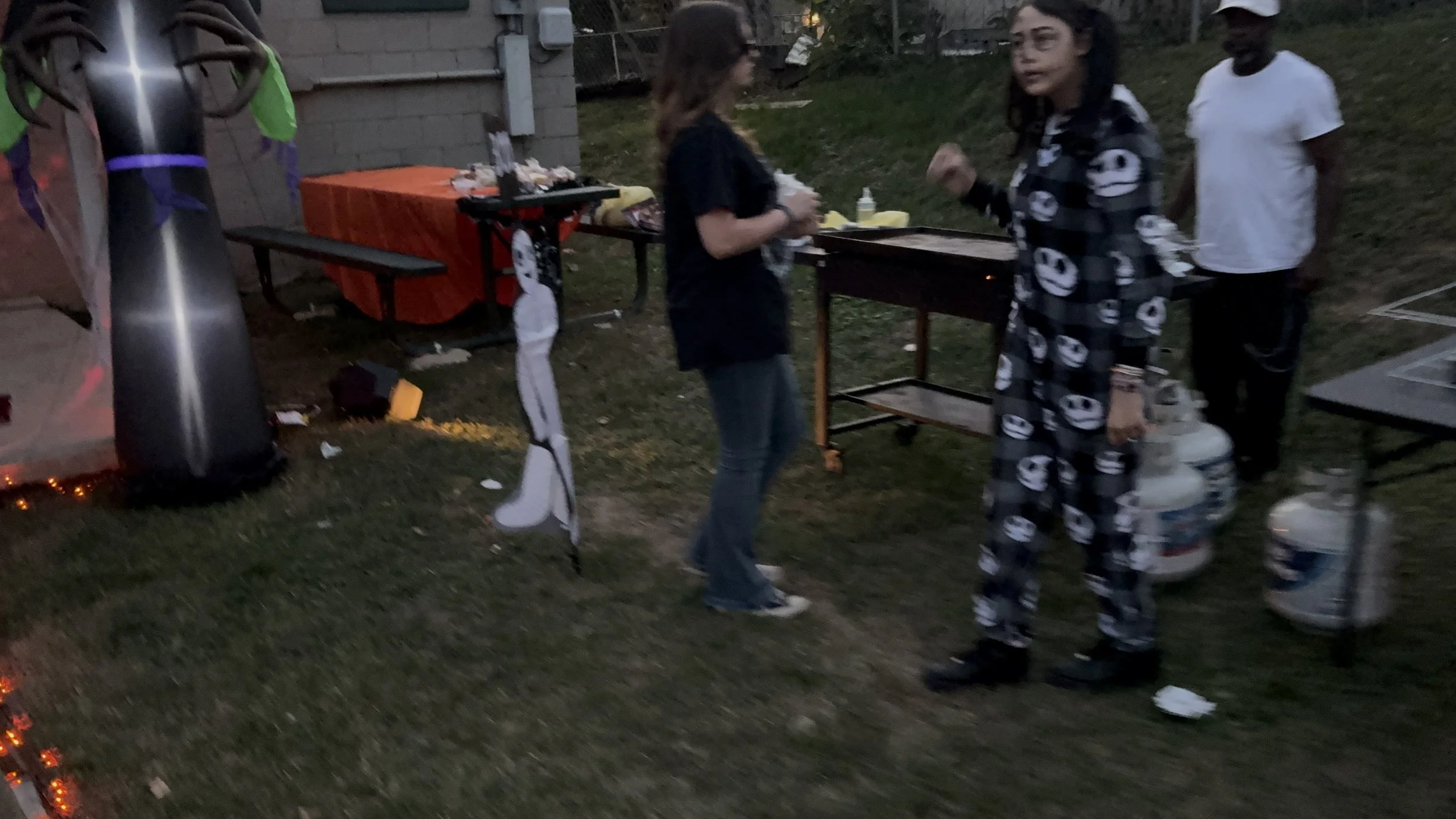 People at an outdoor gathering with a table and propane tanks, some decorations, and a grill in a backyard during dusk.