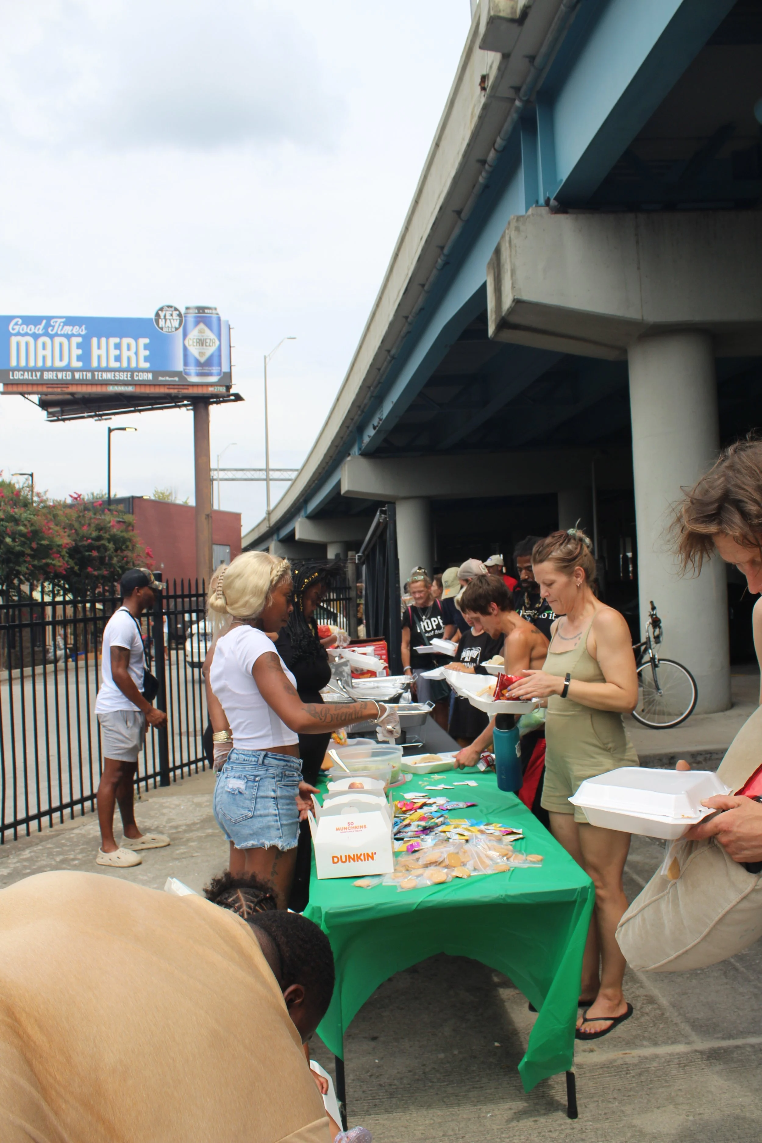 People at a food donation or distribution event under an overpass with a green tablecloth, snacks, and other supplies.