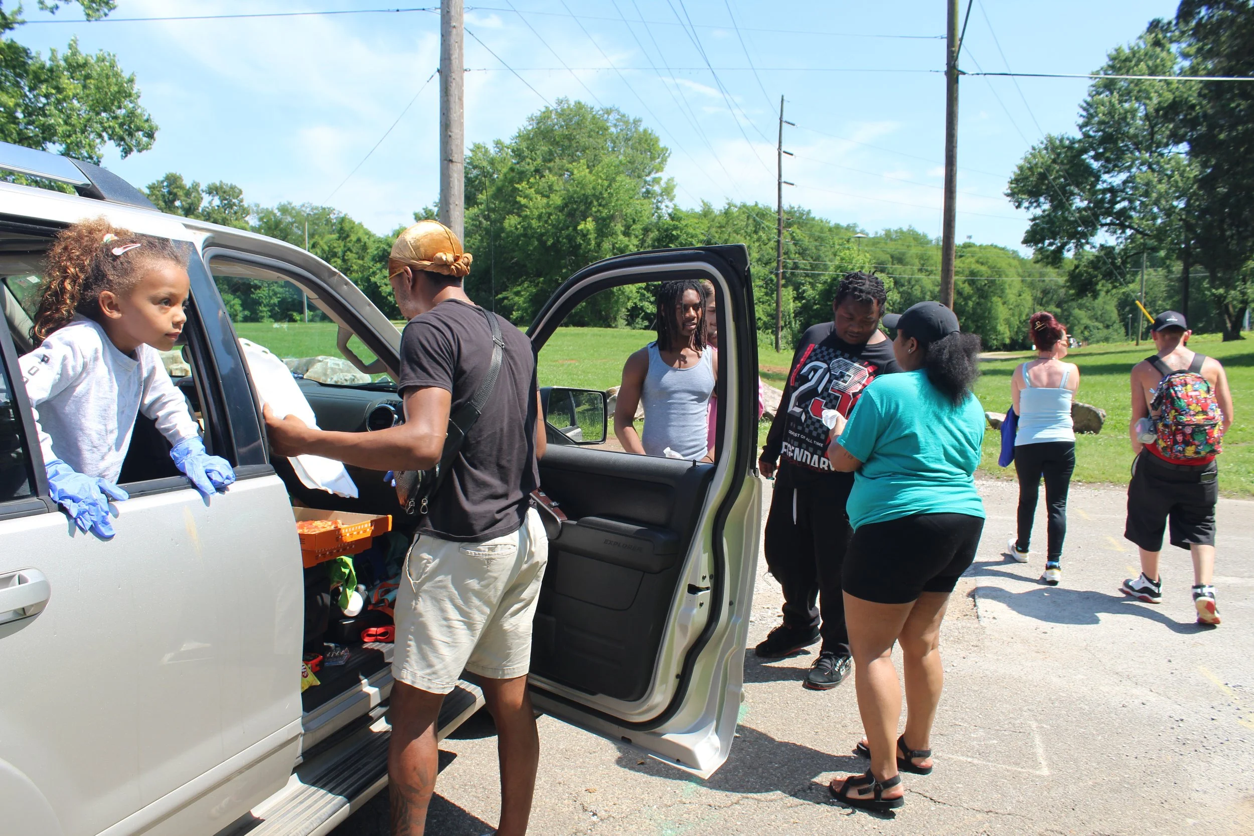 Group of people gathered outside around a white vehicle on a sunny day, with some people talking and a young girl leaning out of the car window.