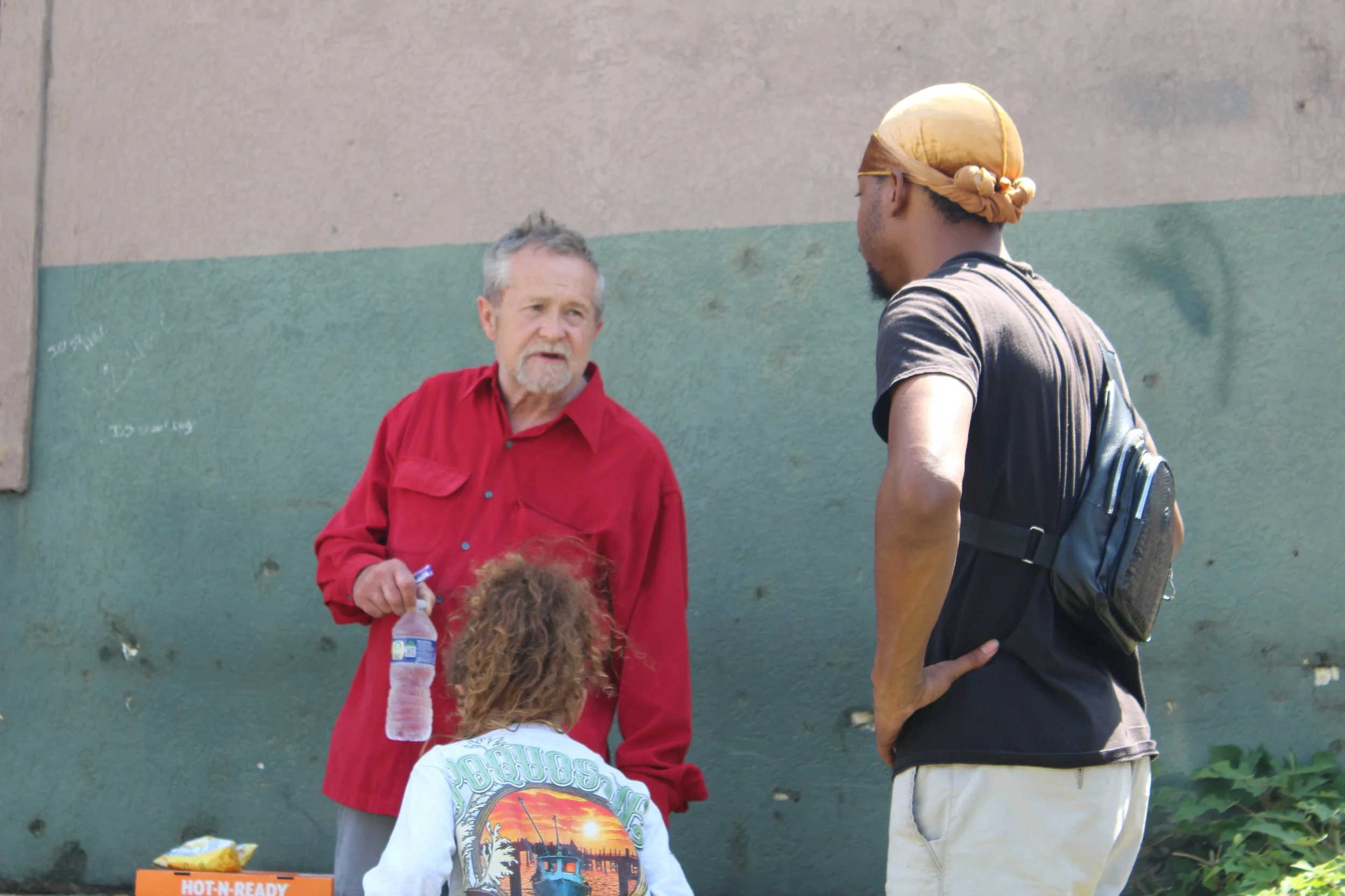 An elderly man in a red shirt speaking to a young man in a black t-shirt and tan cap, with a girl with curly hair facing away from the camera, outdoors against a two-tone wall.