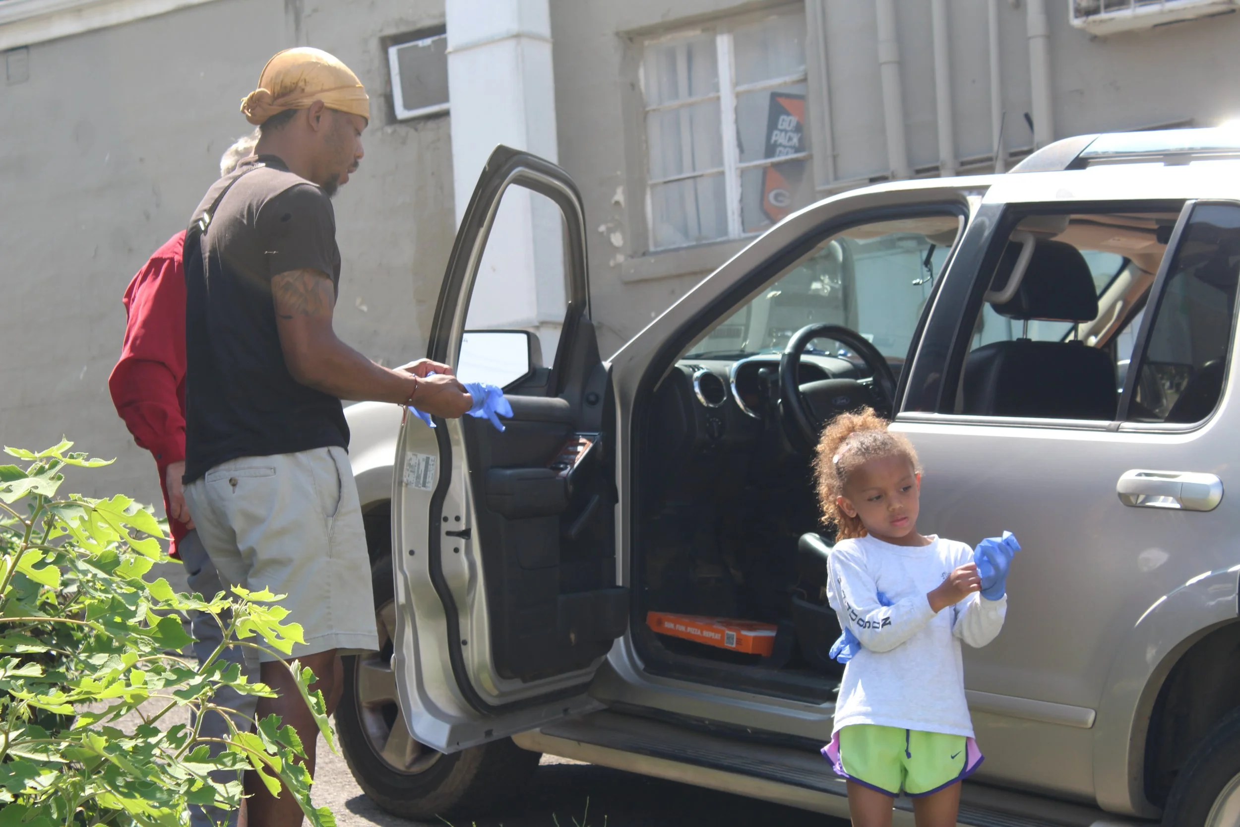A man and a young girl putting on blue gloves next to a silver SUV, with the man standing outside and the girl standing near the open passenger door, in an outdoor urban setting.