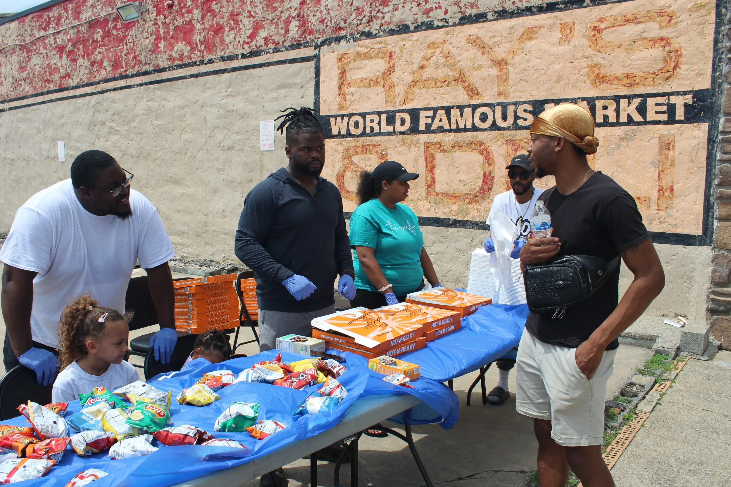 People handing out food and snacks at an outdoor event in front of a beige wall with faded sign that reads 'World Famous Market.'