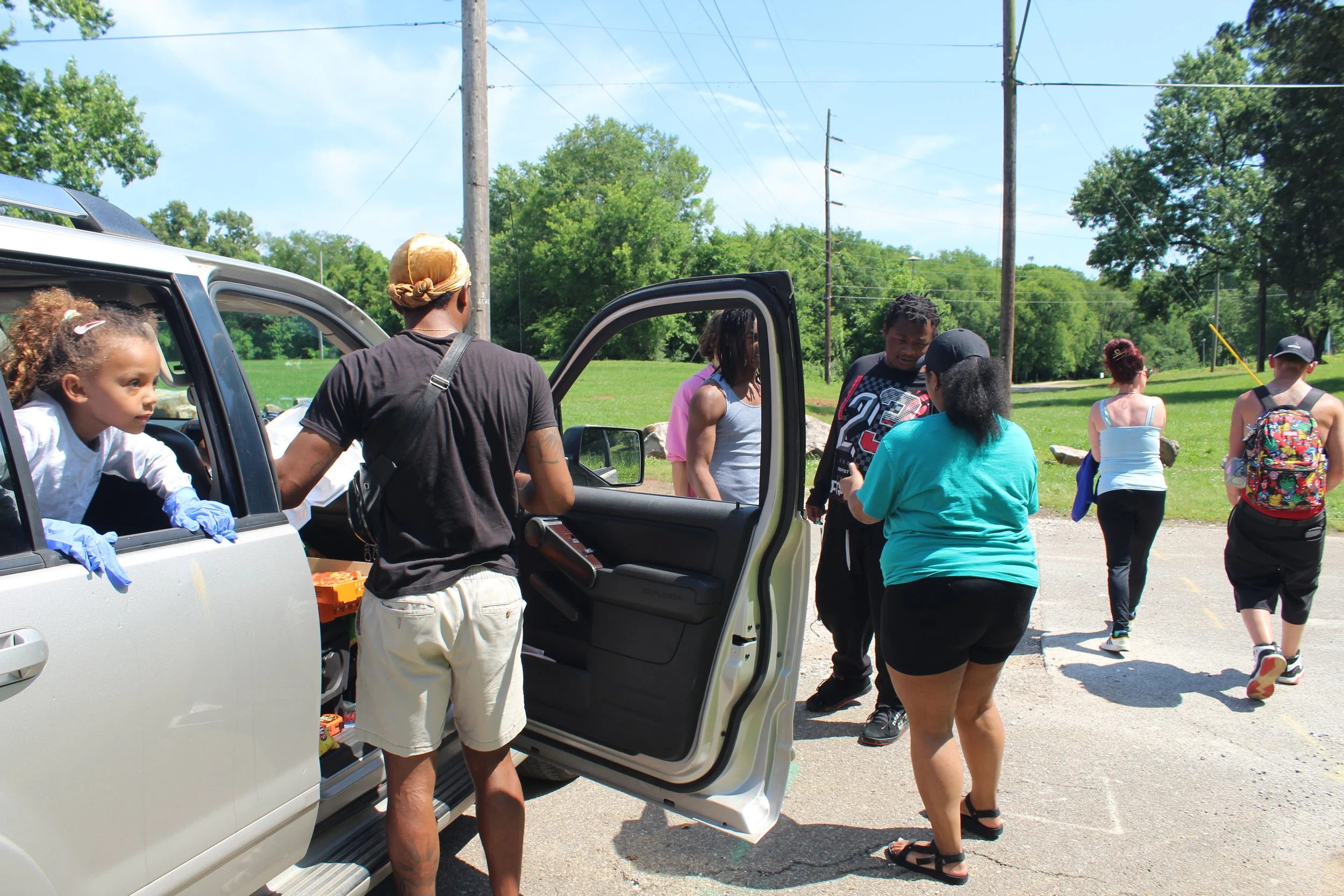 Group of people gathered around an open car door during daytime in a park or outdoor area, some wearing casual clothing and backpacks, with trees and power lines in the background.