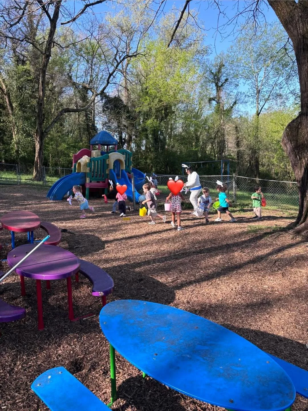 Children in bunny ear headbands playing outdoors in a park near a colorful playground with trees and a blue sky in the background.