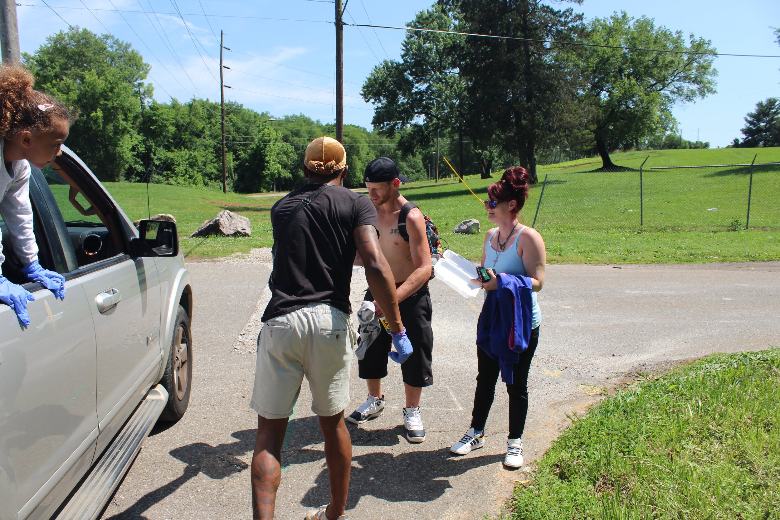 Group of people standing on a parking lot near a grassy field with trees, engaged in conversation with one person leaning out of a vehicle window. One person holds a container and another holds a phone, with blue gloves on some individuals' hands.