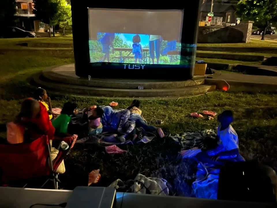 Group of children and adults watching a movie outdoors on a large screen in a park at night, with some sitting on blankets and others on the grass.