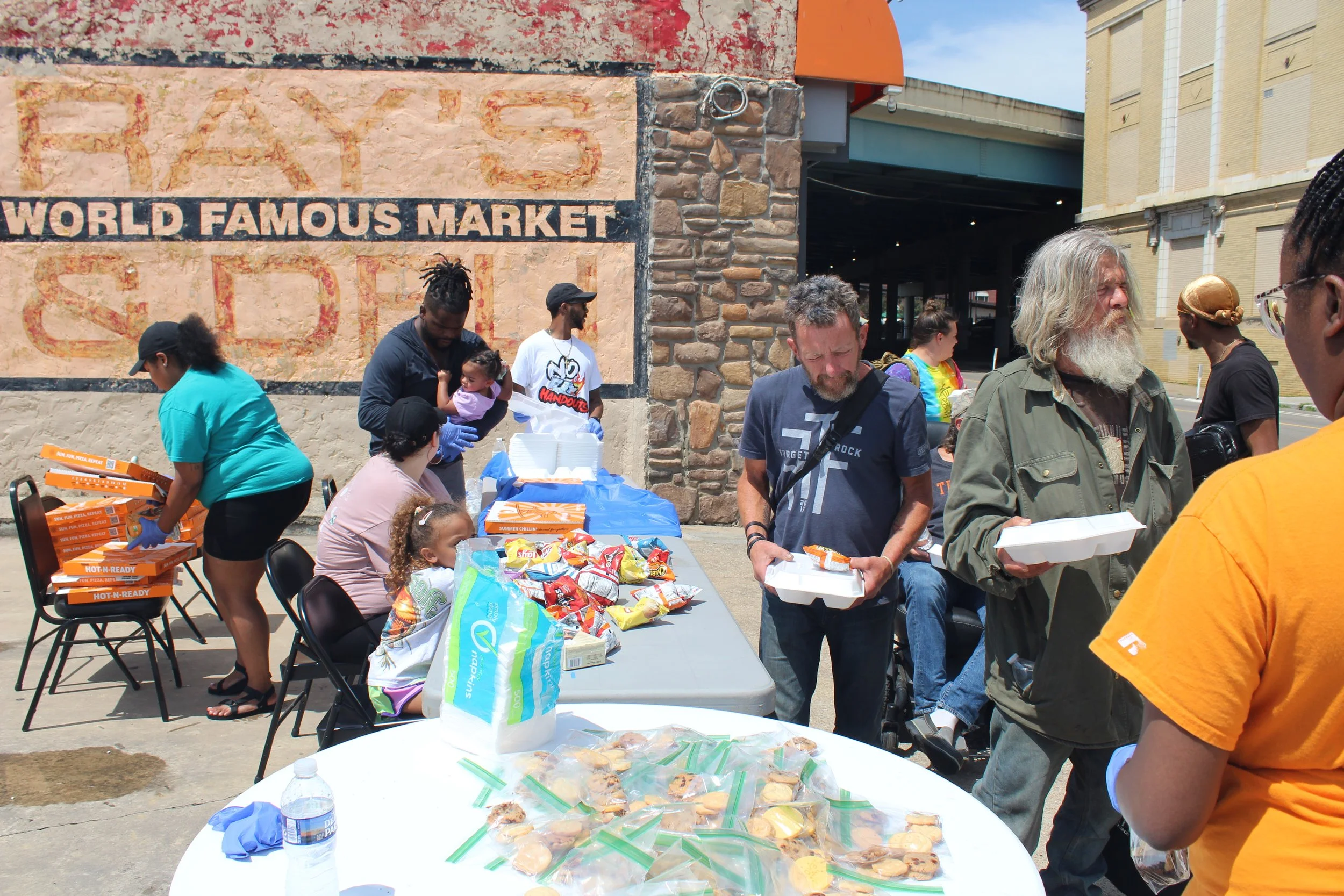 People standing in line at a food distribution table outside a market, with boxes of food and packaged baked goods on the table, and a sign that reads 'World Famous Market' on the wall behind them.