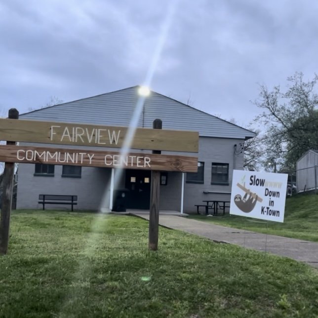 A sign in front of a two-story community center building labeled 'Fairview Community Center,' with a smaller sign reading 'Slow Down in K-Town' featuring a turtle illustration, green grass, and a paved walkway.