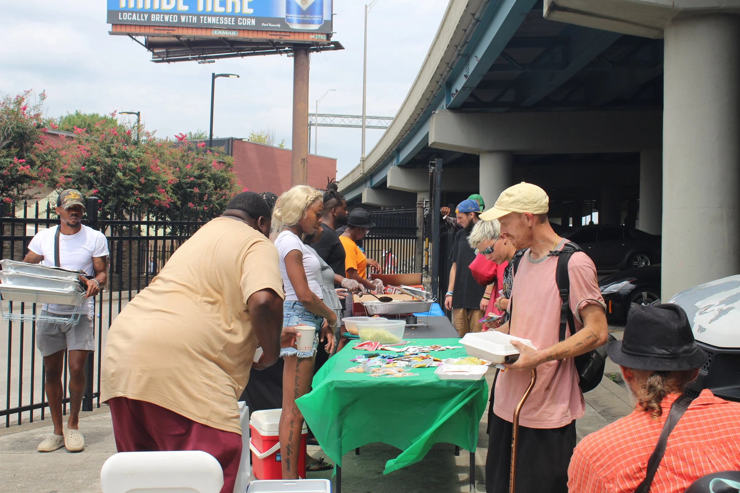 People standing in line at an outdoor food booth under a bridge. The table has a green tablecloth with various food items and supplies. A man in a pink shirt and a beige cap is holding a food container. Several other people are waiting or being serve