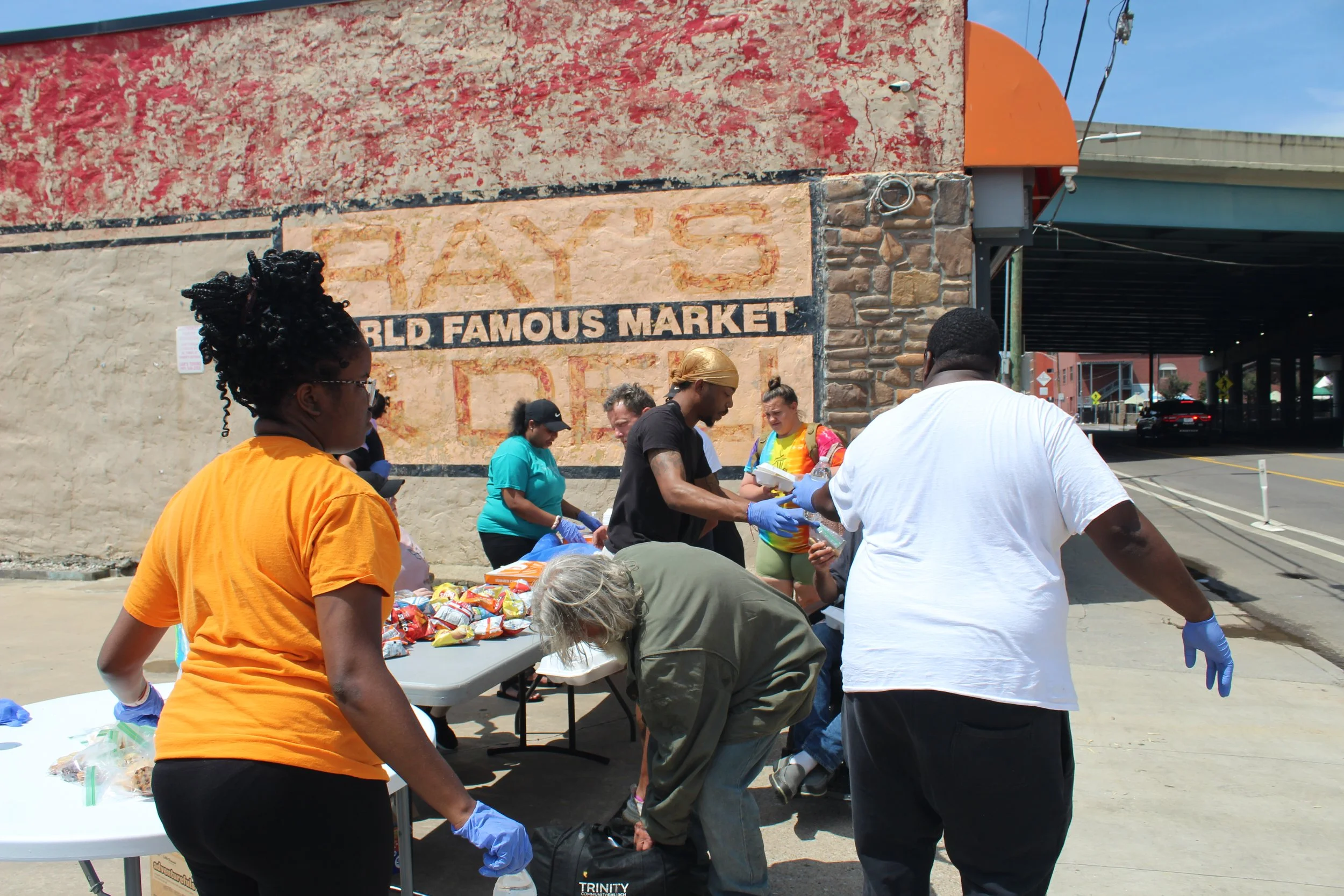 People distributing food and supplies at a community event outside a building with a sign that reads 'FAY'S WORLD FAMOUS MARKET.' Some individuals are wearing gloves and handling food items on tables.