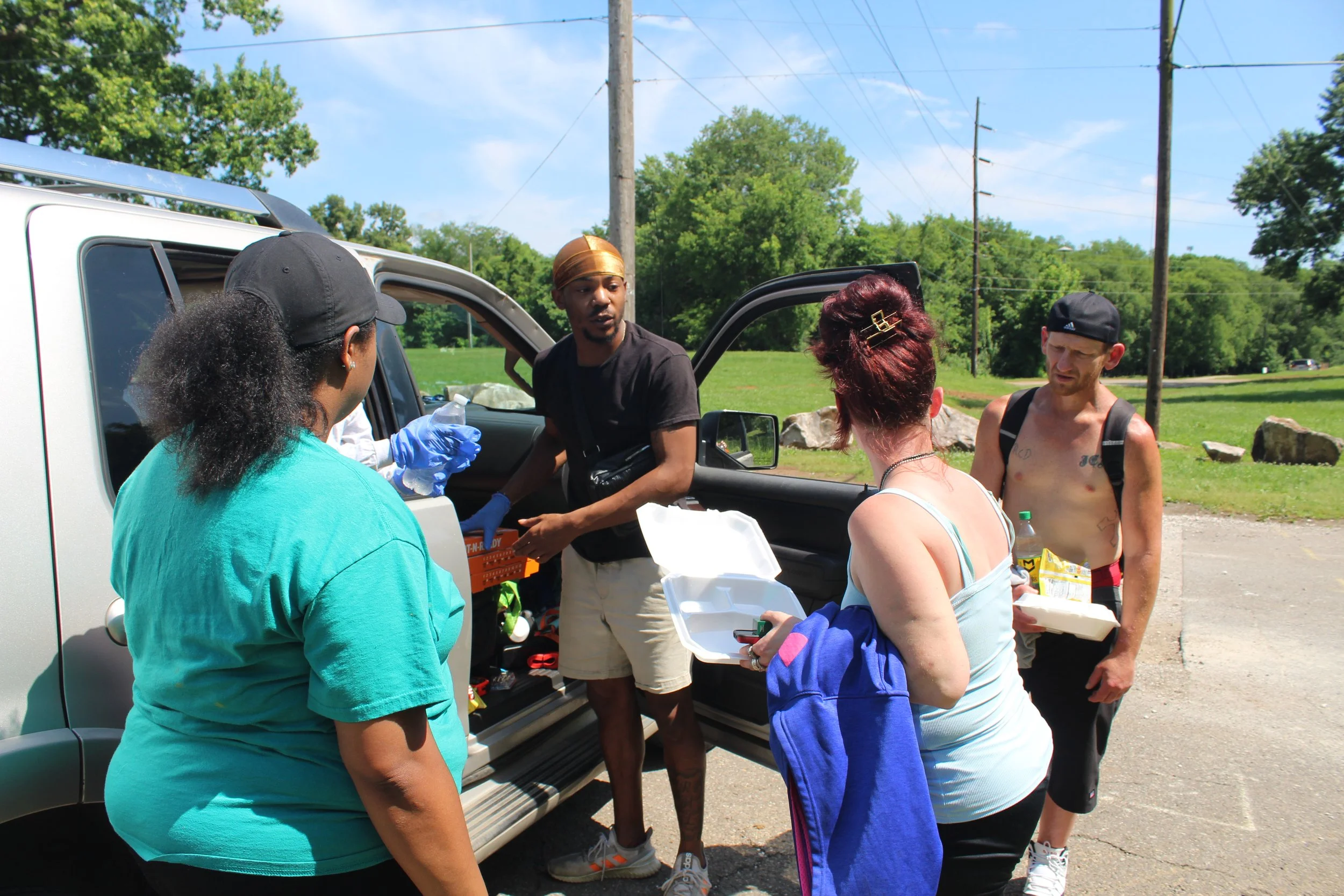 A group of people gathering around an open vehicle in a park-like setting, with green trees, rocks, and power lines in the background on a sunny day.