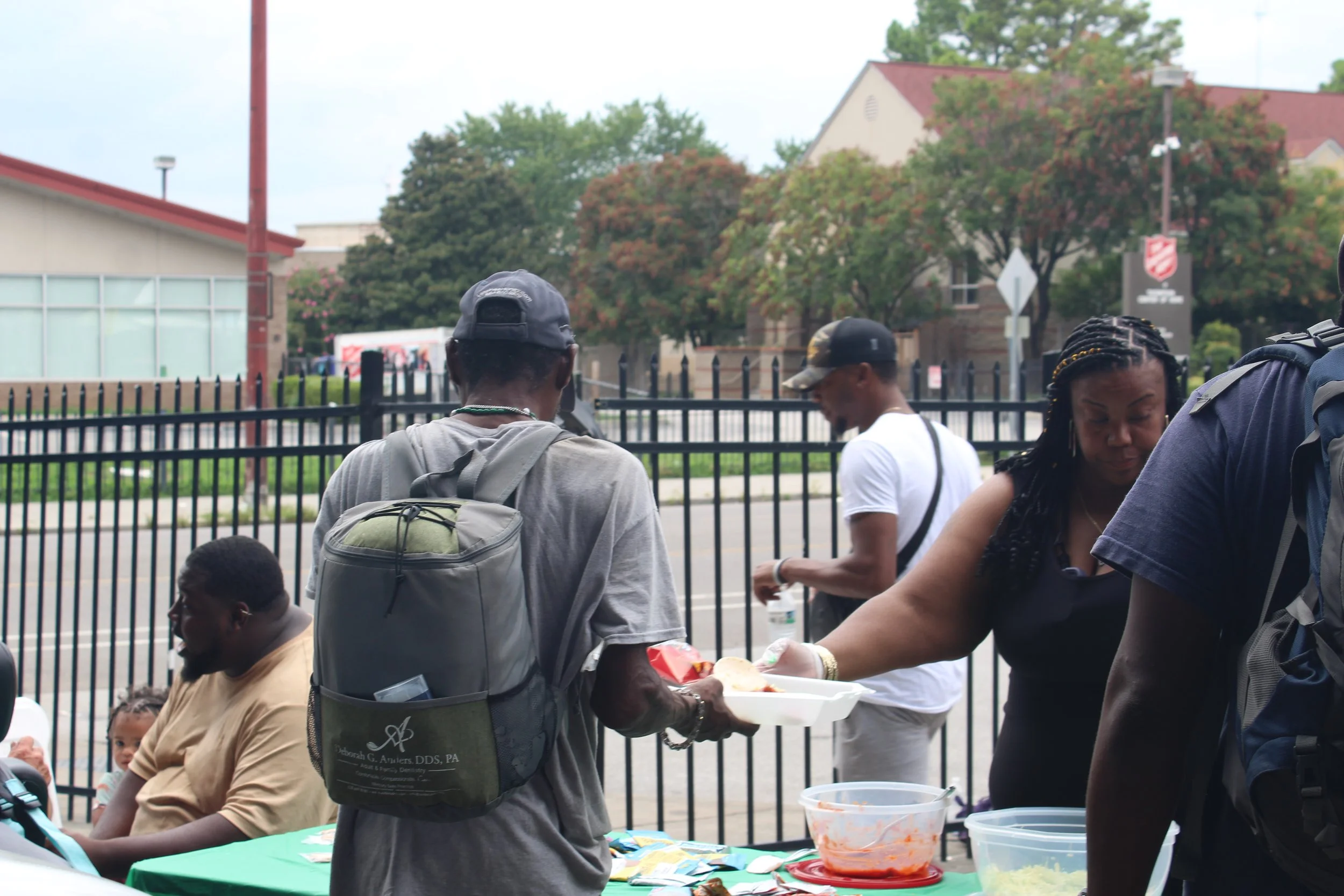 People gathered outdoors at a community event, with some serving food and others sitting at a table, near a black metal fence and brick buildings in the background.