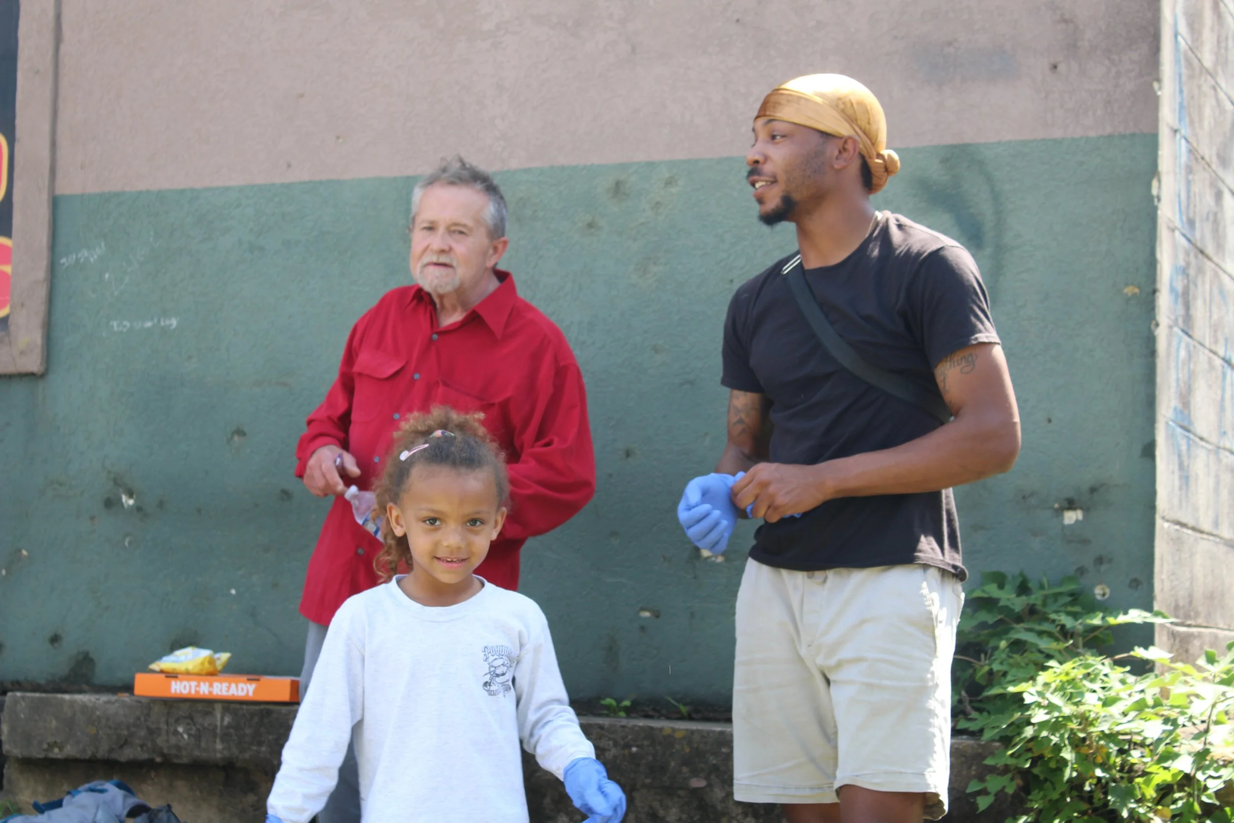 A young girl smiling in front of two men, one older with gray hair and beard wearing a red shirt, and the other younger with a beard and bandana, wearing a black shirt and gloves, outdoors in a neighborhood.