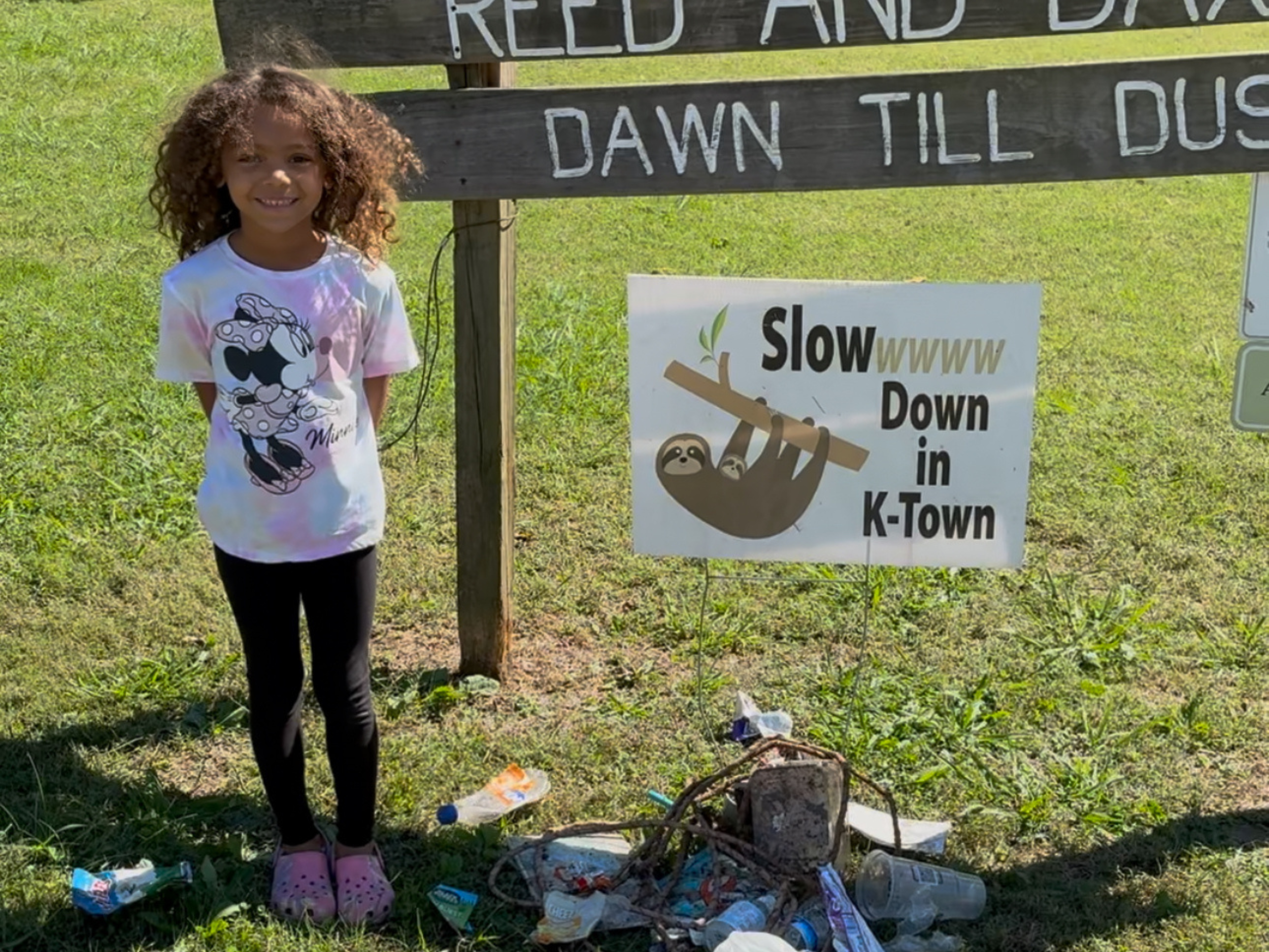 A young girl with curly hair, wearing a Minnie Mouse t-shirt and black leggings, standing outdoors on grass near a sign that says 'Slow Down in K-Town' with a picture of a sloth. The ground around her has trash and bottles.