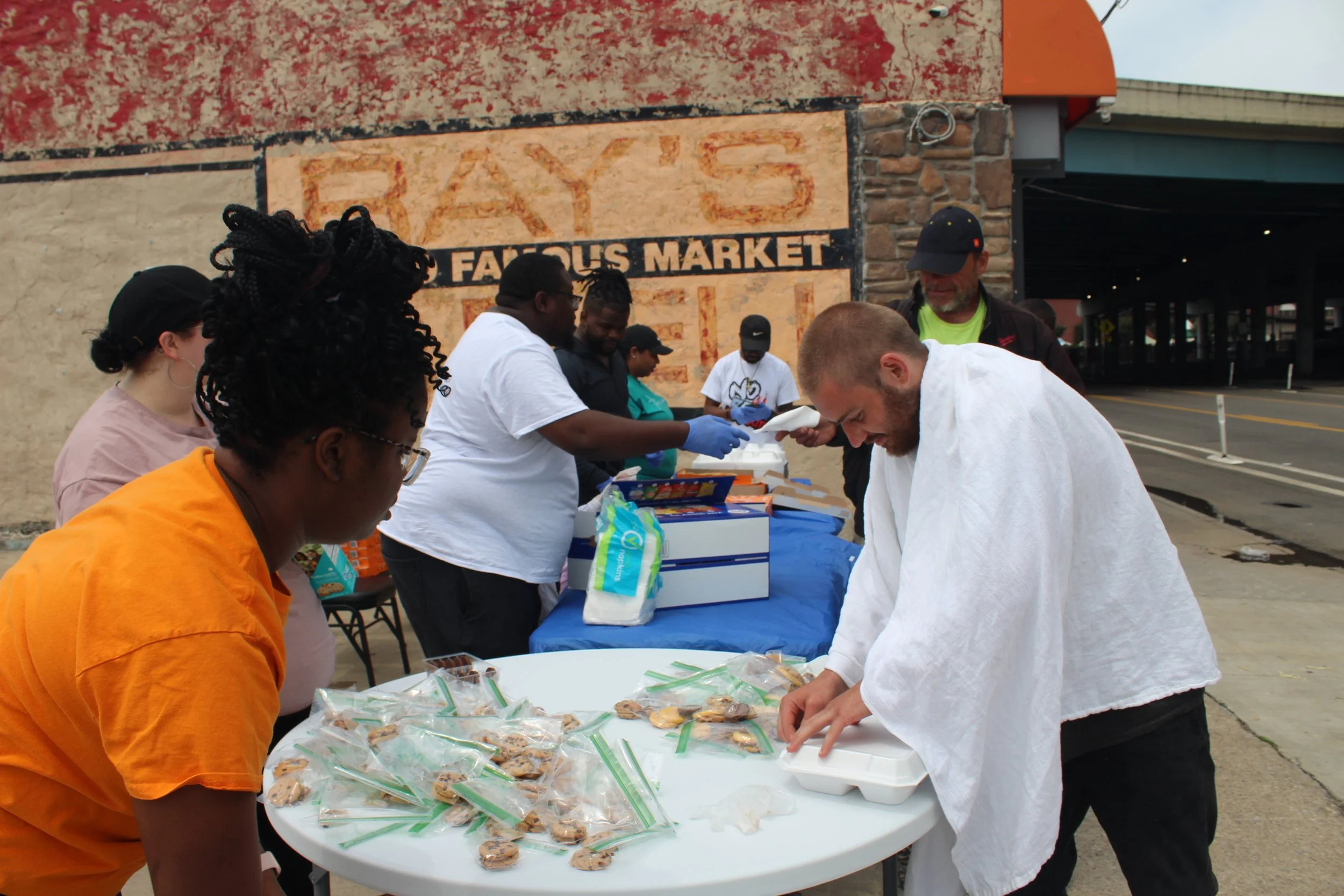 People distributing cookies at a table outside a market, with a large sign reading 'Famous Market' in the background.