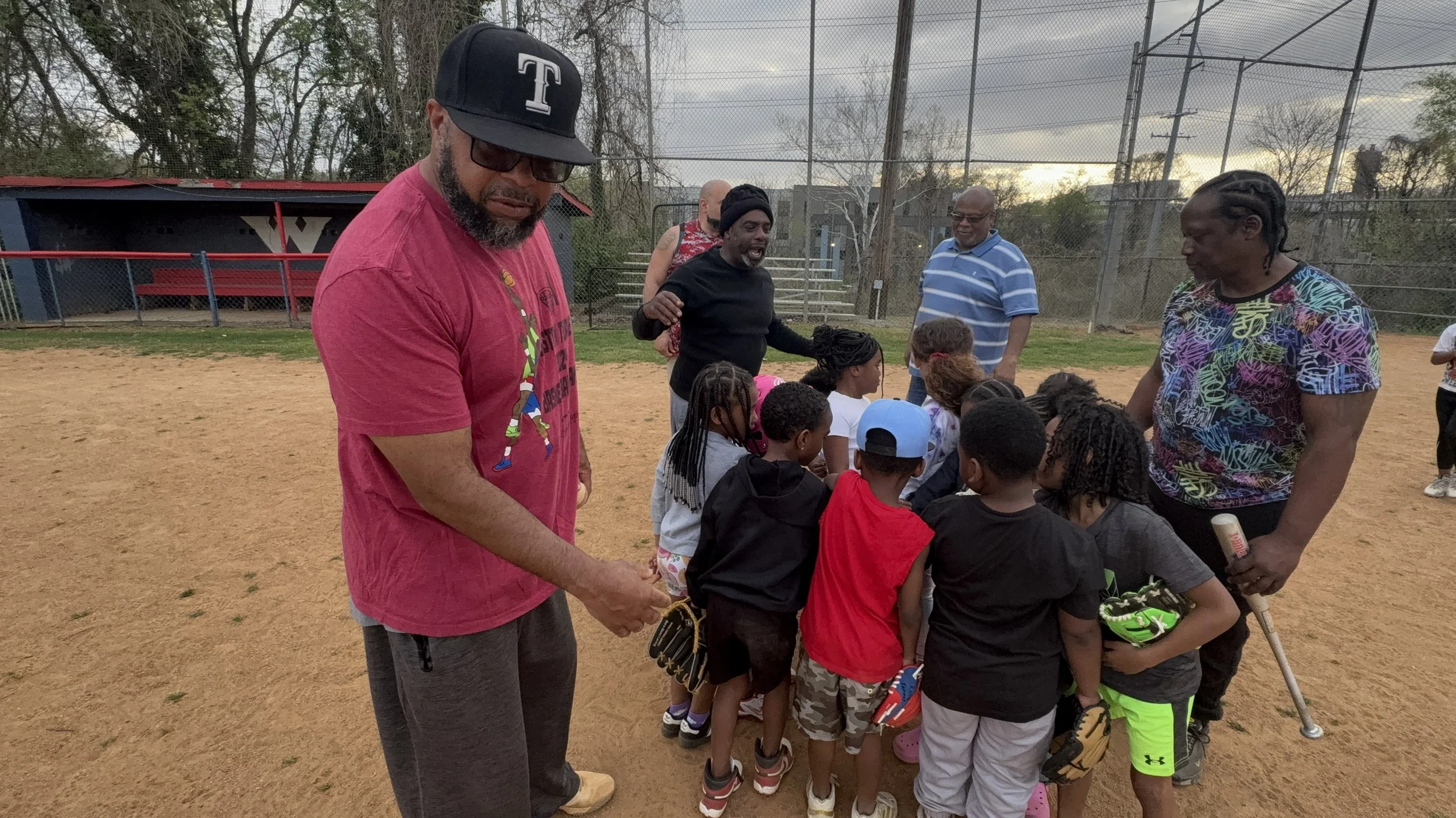 A group of children and adults gathered on a baseball field, with some holding baseball gear, engaged in a discussion or activity, with a baseball backstop and trees in the background.