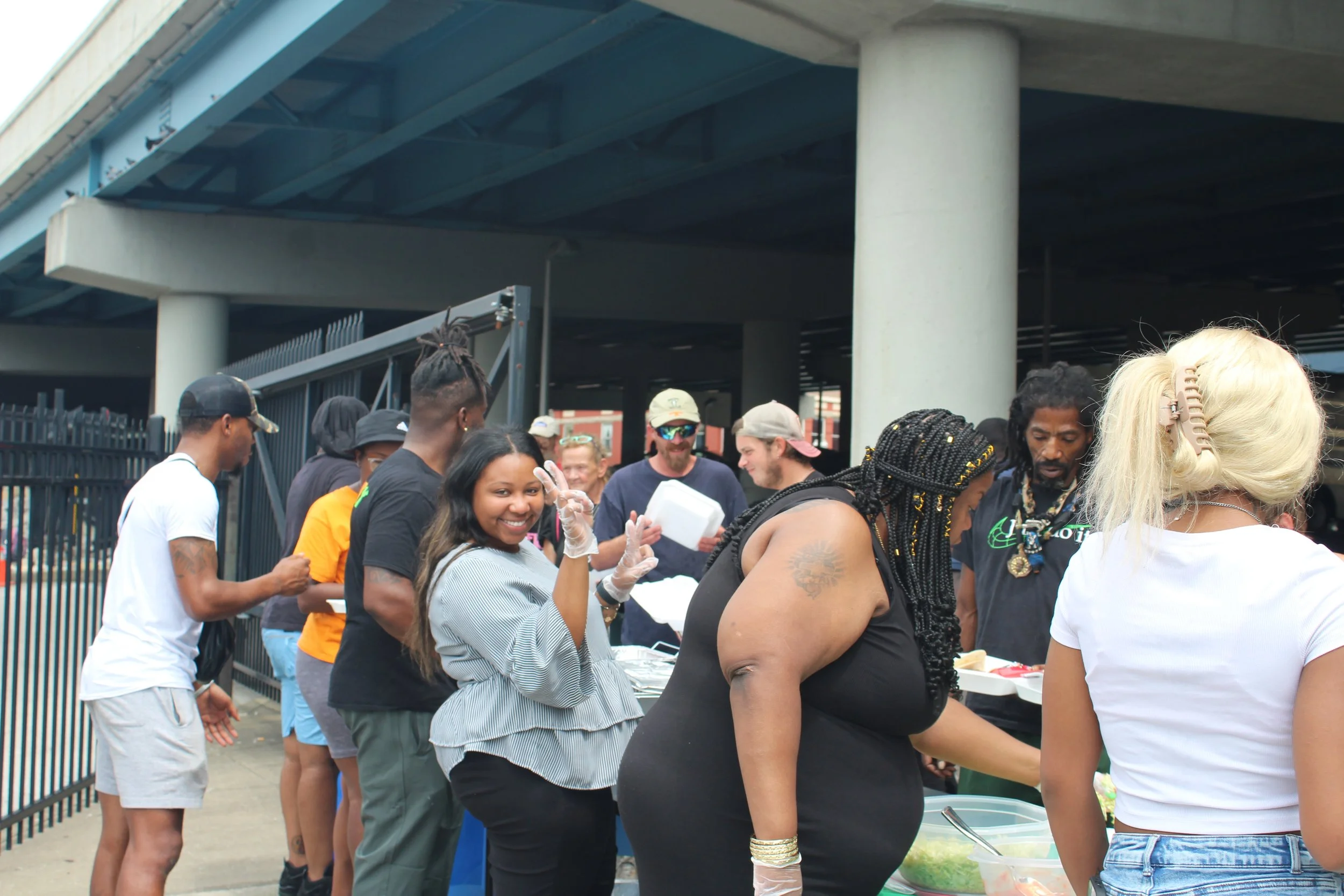 People standing in line at an outdoor food or drink stand under an overpass, with some people smiling and making peace signs, and others serving or waiting for their orders.