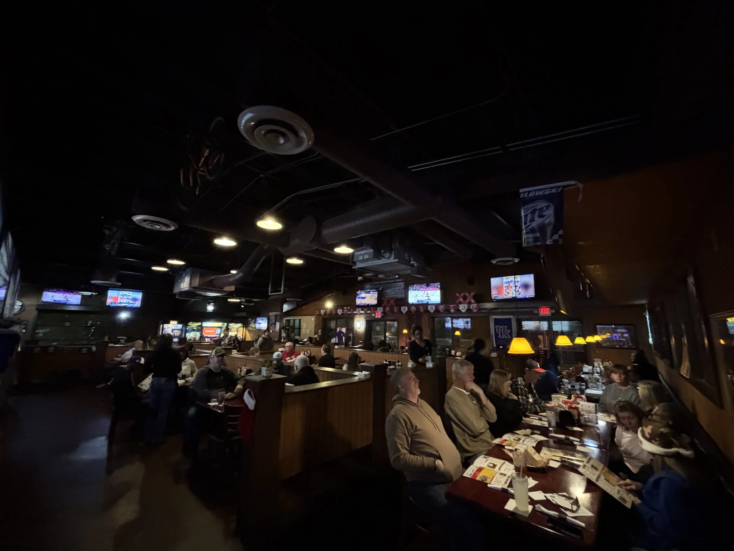 A crowded indoor restaurant or bar area with people sitting at tables, looking at menus, and socializing. There are multiple television screens on the walls displaying sports games, and the ceiling has exposed ducts and circular lighting fixtures.