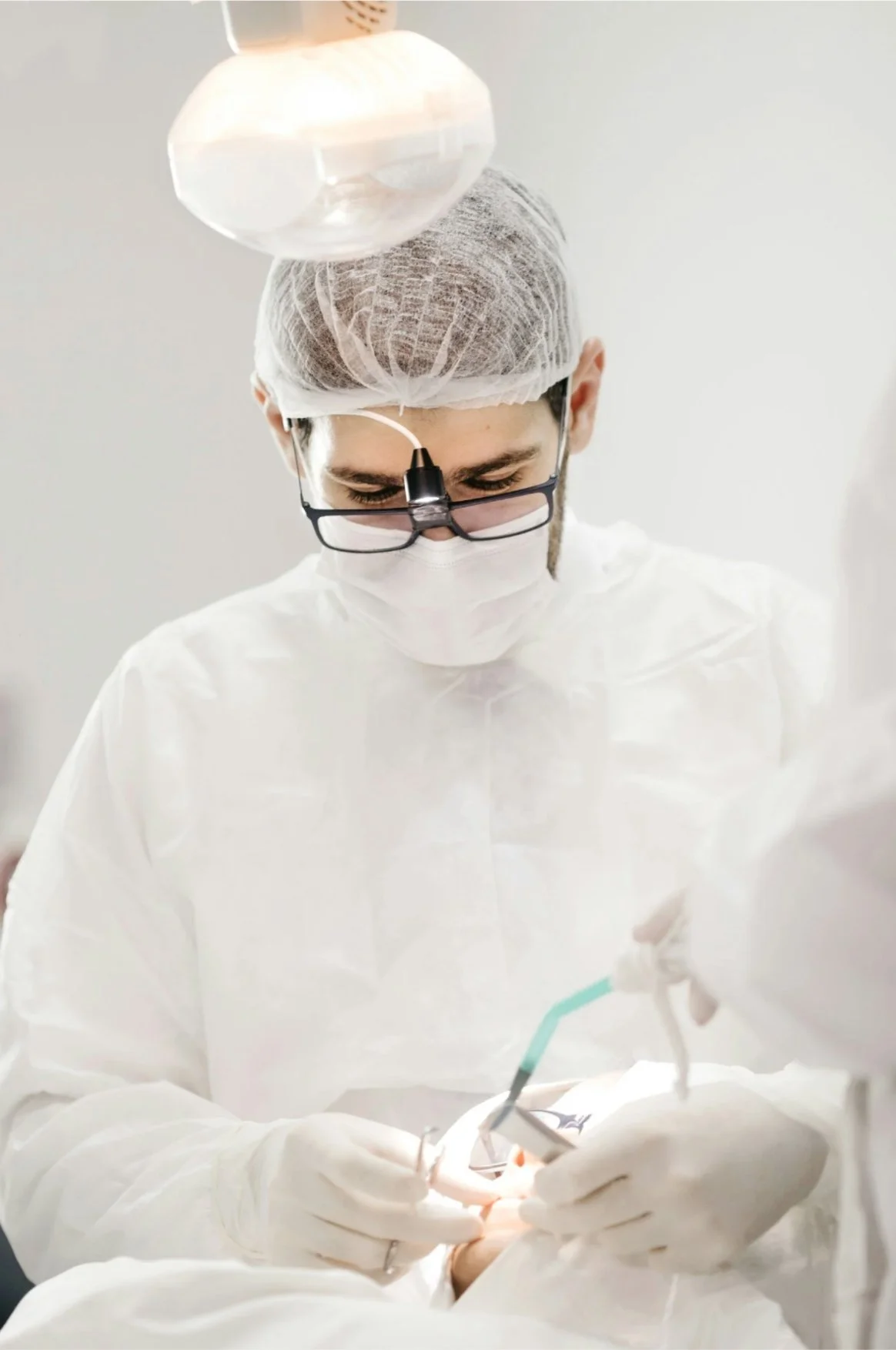 A male surgeon wearing a hairnet, glasses with a light, a face mask, and sterile gloves performing surgery in a medical setting.