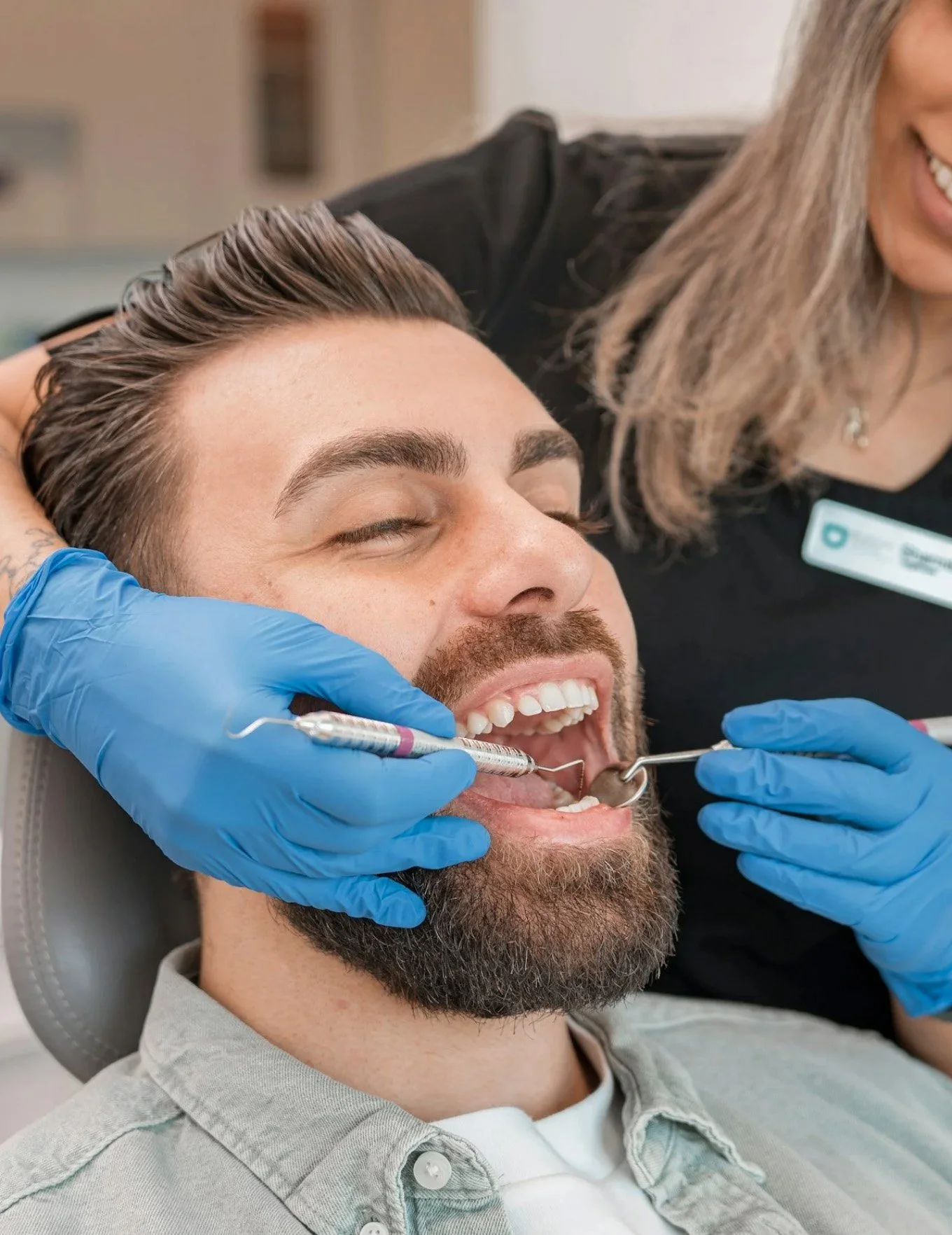 A man with a beard getting dental treatment from a dental professional wearing blue gloves. The man is smiling with his mouth open, and the dental professional is using dental tools.