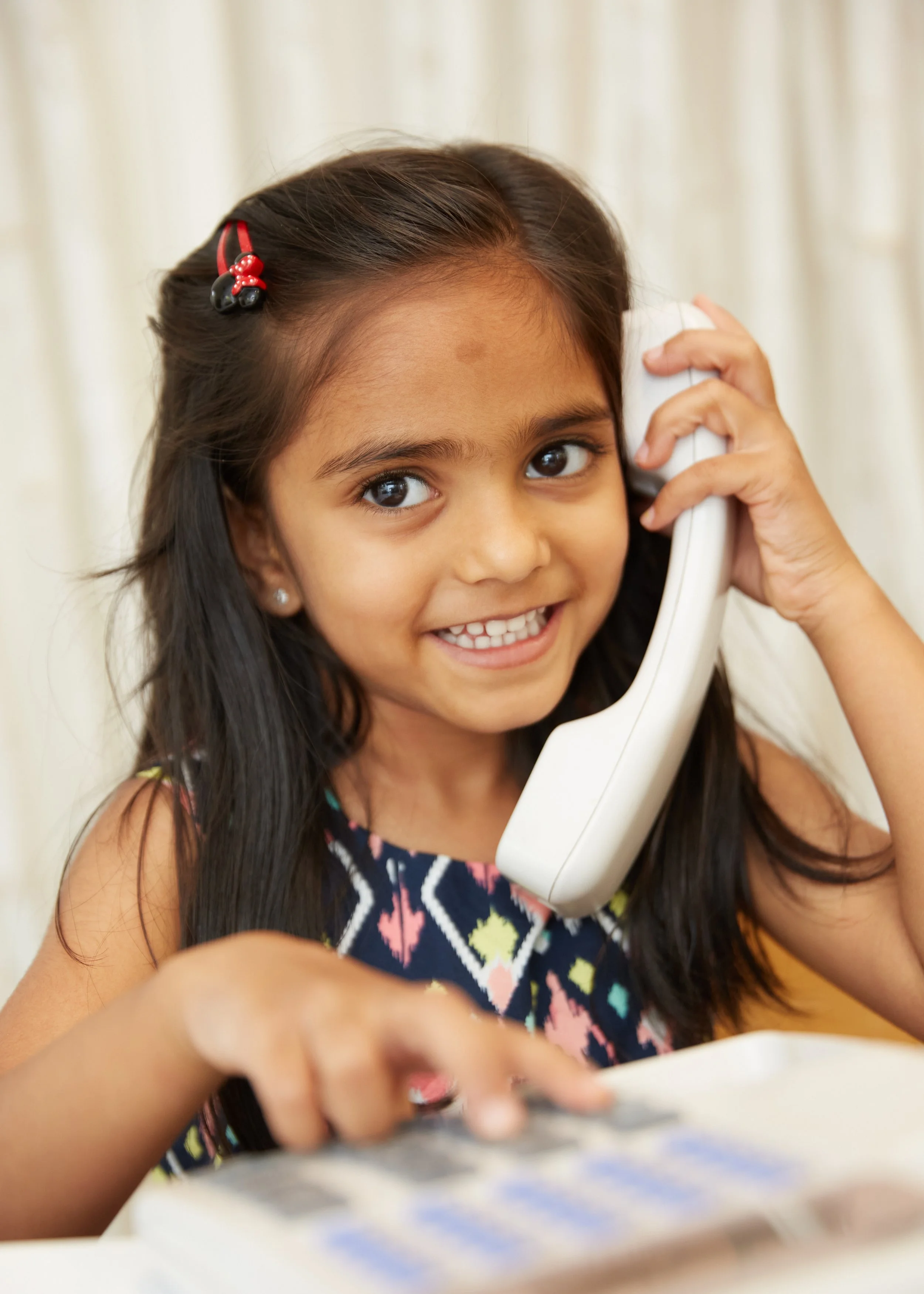 A young girl with dark hair, wearing earrings and a colorful sleeveless top, is smiling and talking on a white landline telephone, with a blurred background.