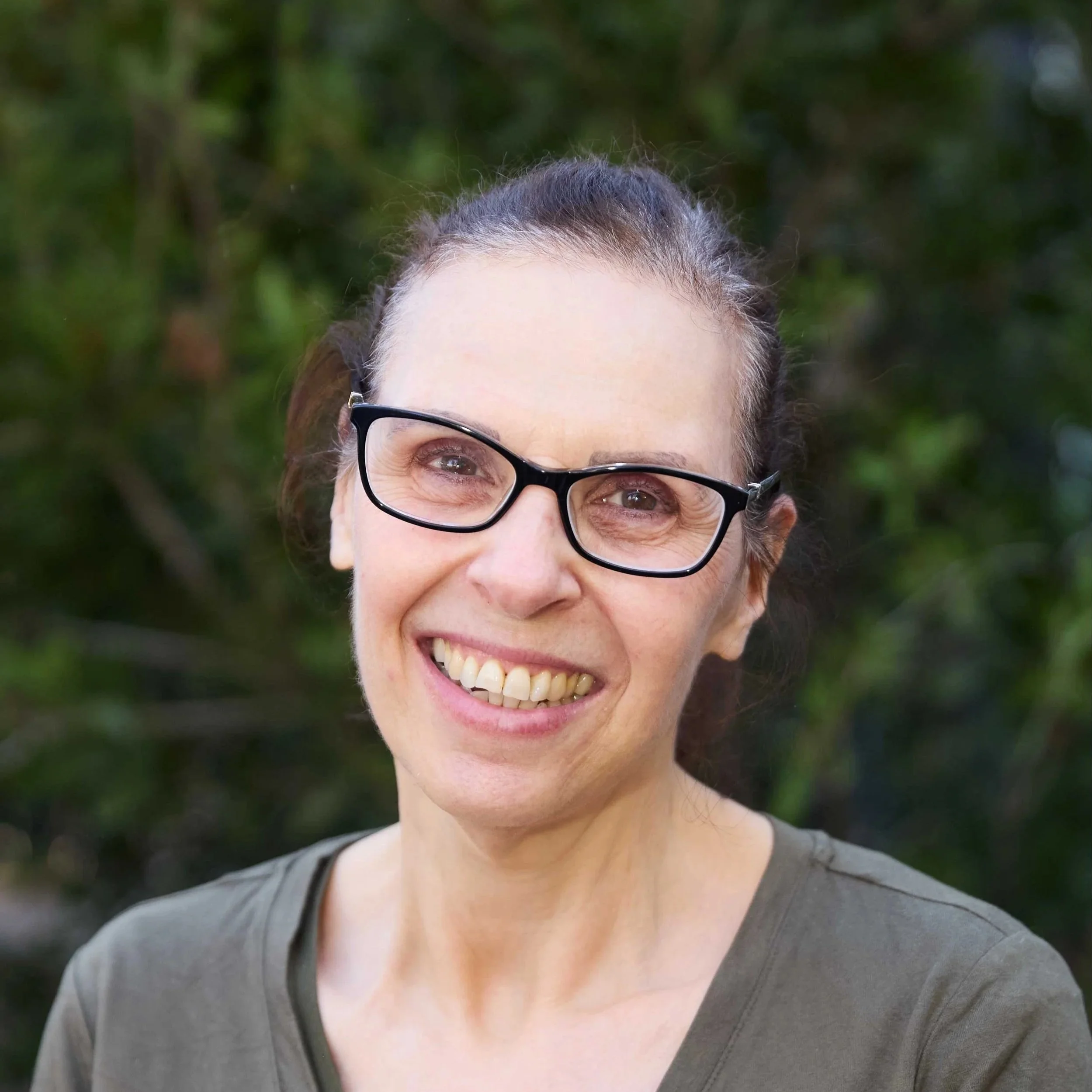 A woman smiling in front of a brick wall, wearing glasses, a black jacket, and a colorful scarf.