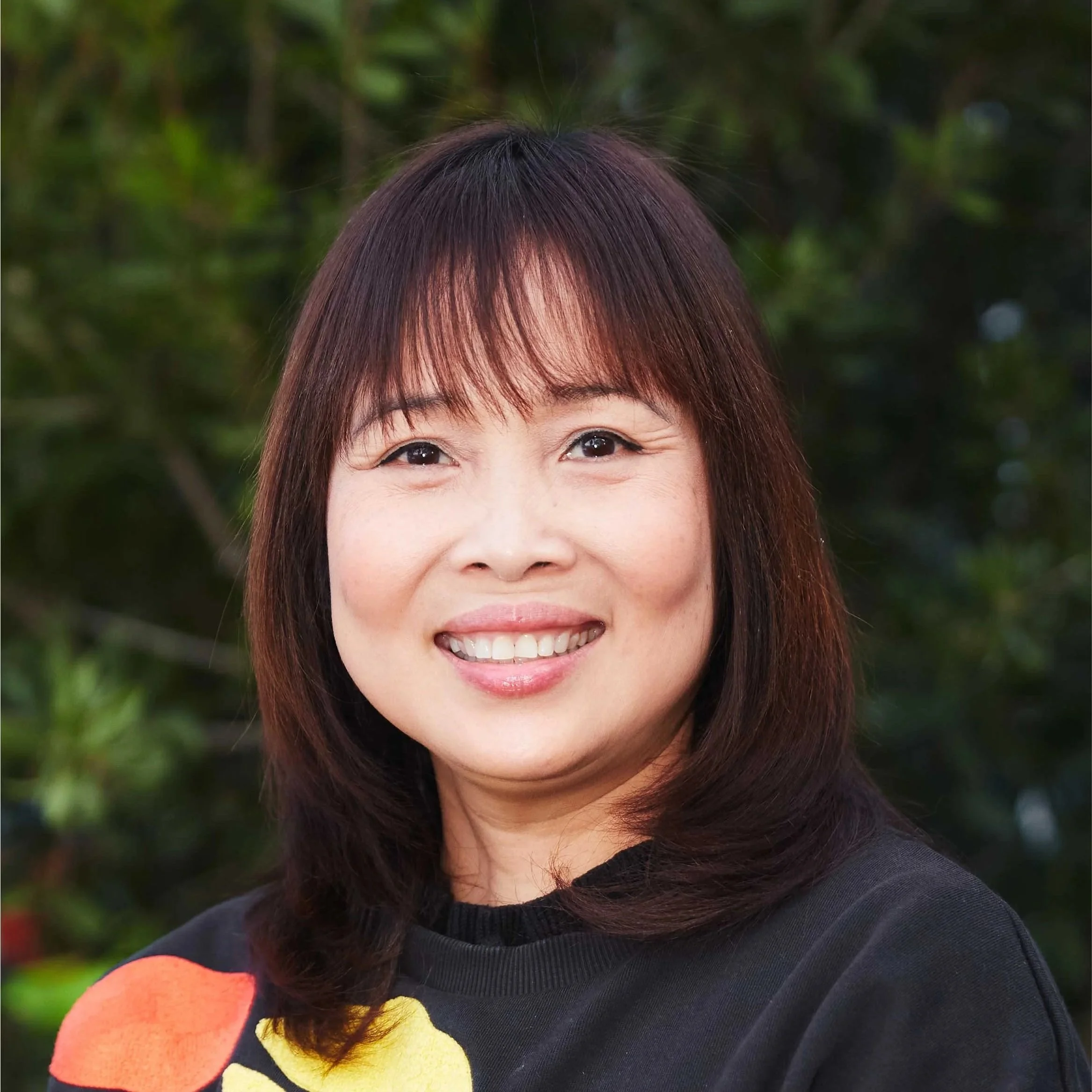 Close-up of a smiling Asian woman with dark hair pulled back, wearing a black quilted jacket, standing in front of a brick wall.