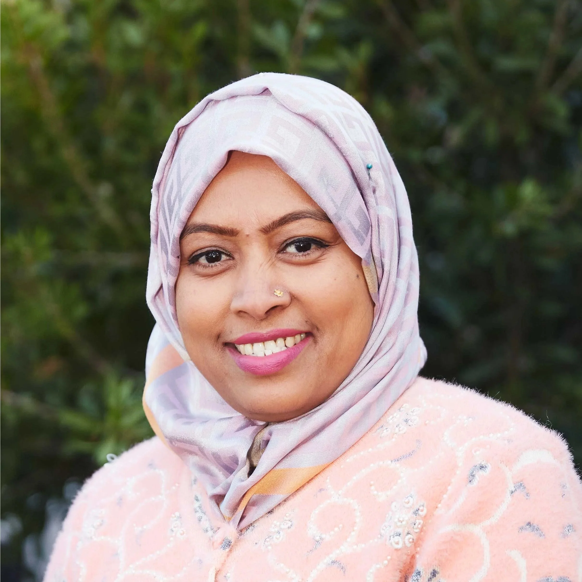 A woman wearing a black headscarf and a blue polka dot top, standing in front of a backdrop of light-colored wooden logs.