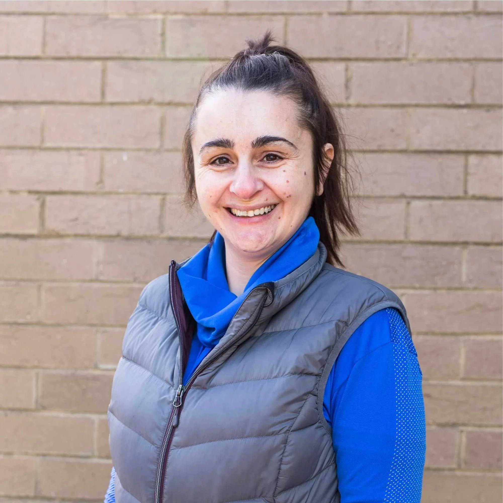 A woman smiling outdoors against a brown brick wall, wearing a gray vest over a blue jacket.