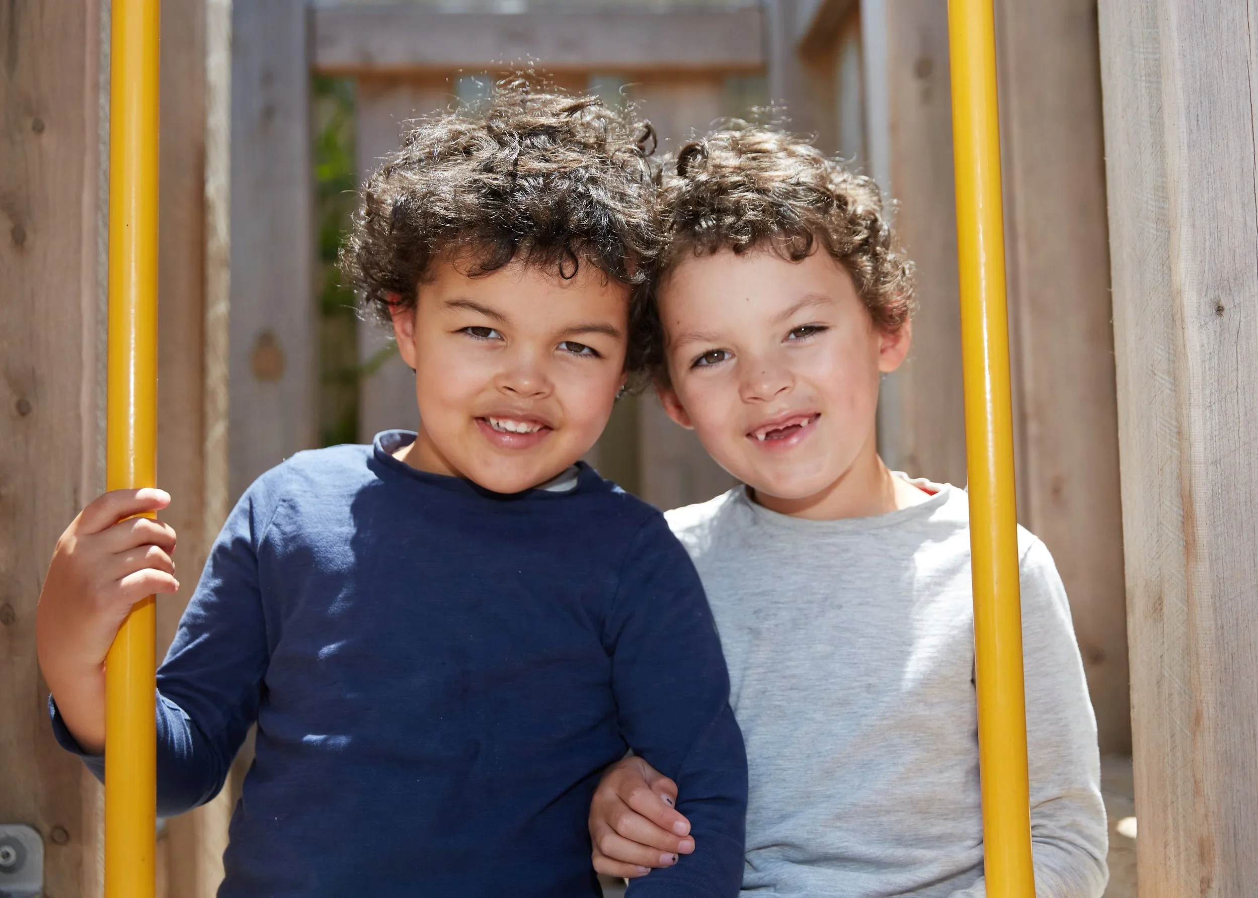 Two young boys with curly hair smiling and standing close together on a playground, holding onto yellow bars.
